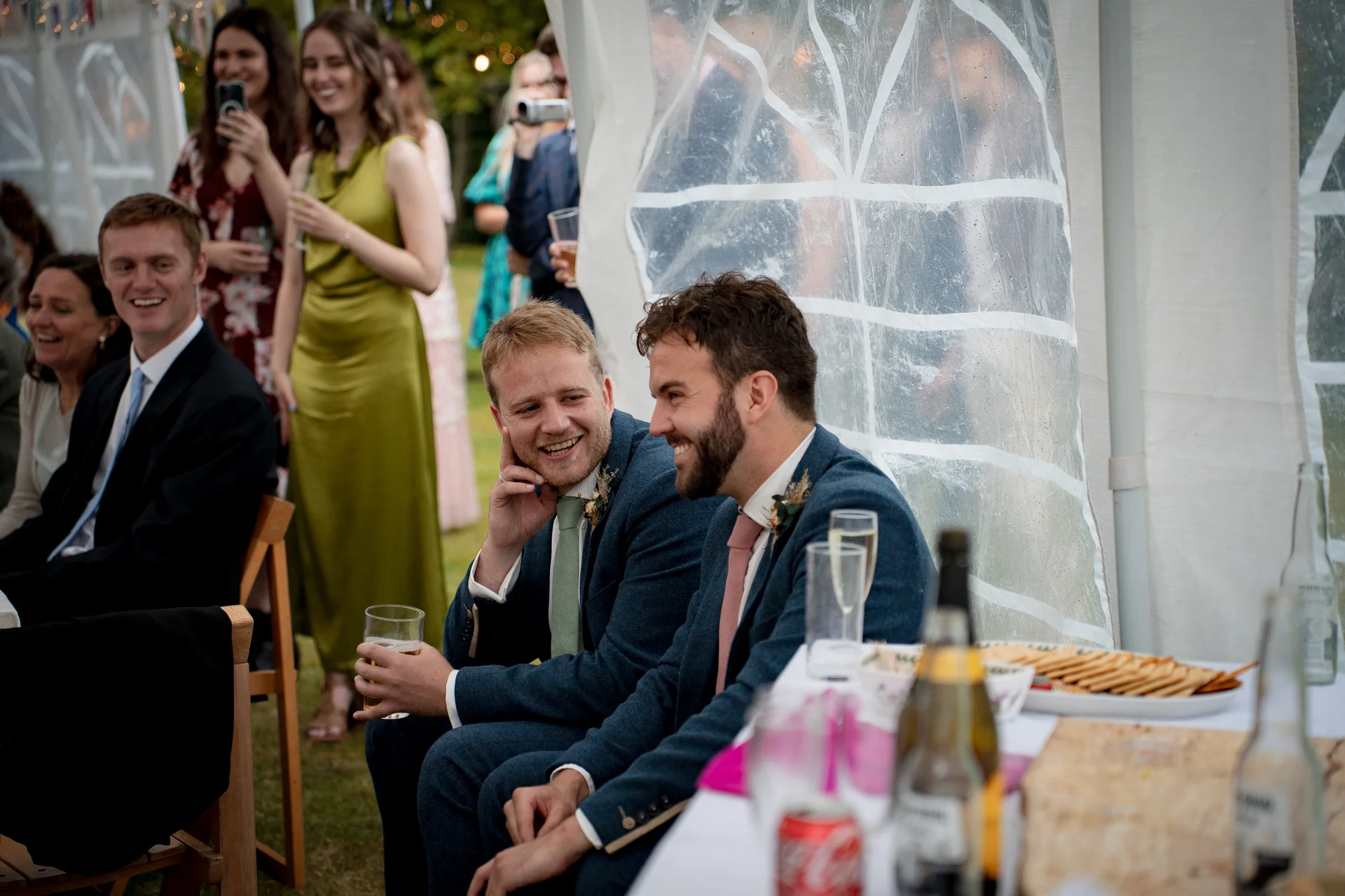 Two grooms share a joyful laugh while seated together at a marquee wedding reception alongside smiling guests.