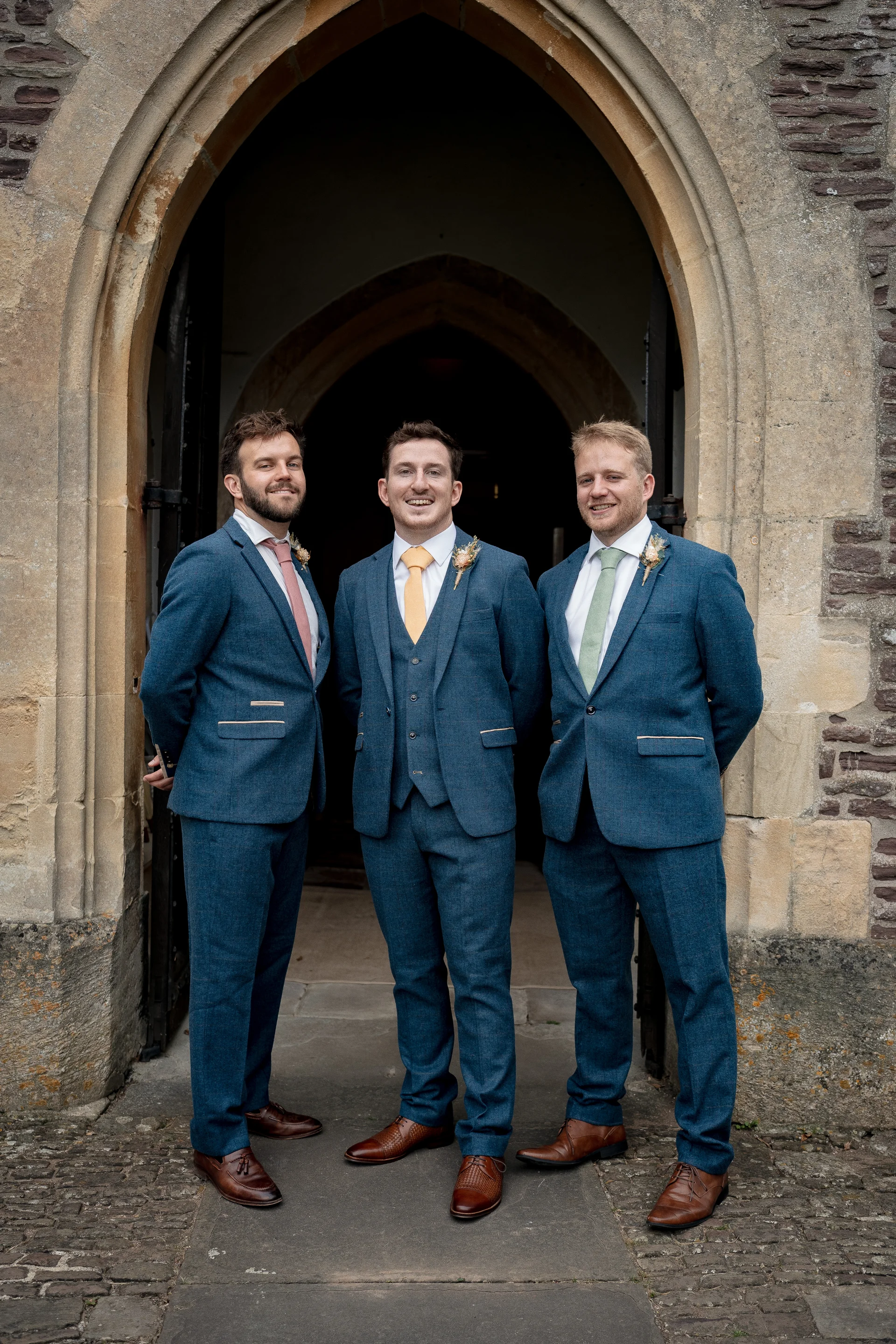 Three smiling groomsmen wearing matching blue suits and distinct pastel ties pose together framed inside a historic stone church doorway.