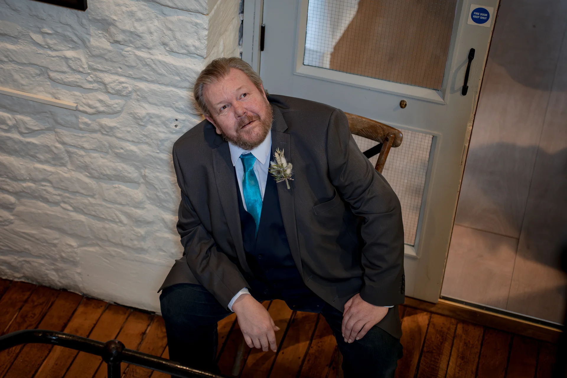 A solo groom in a charcoal suit and bright turquoise tie sits on a wooden chair against a white whitewashed brick wall.