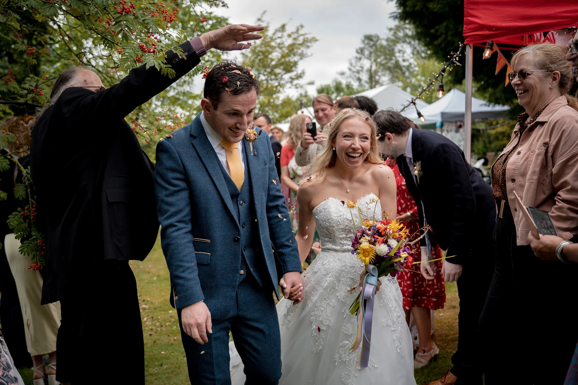 A joyful newlywed couple walks hand in hand through cheering guests who shower them with confetti during an outdoor wedding celebration.