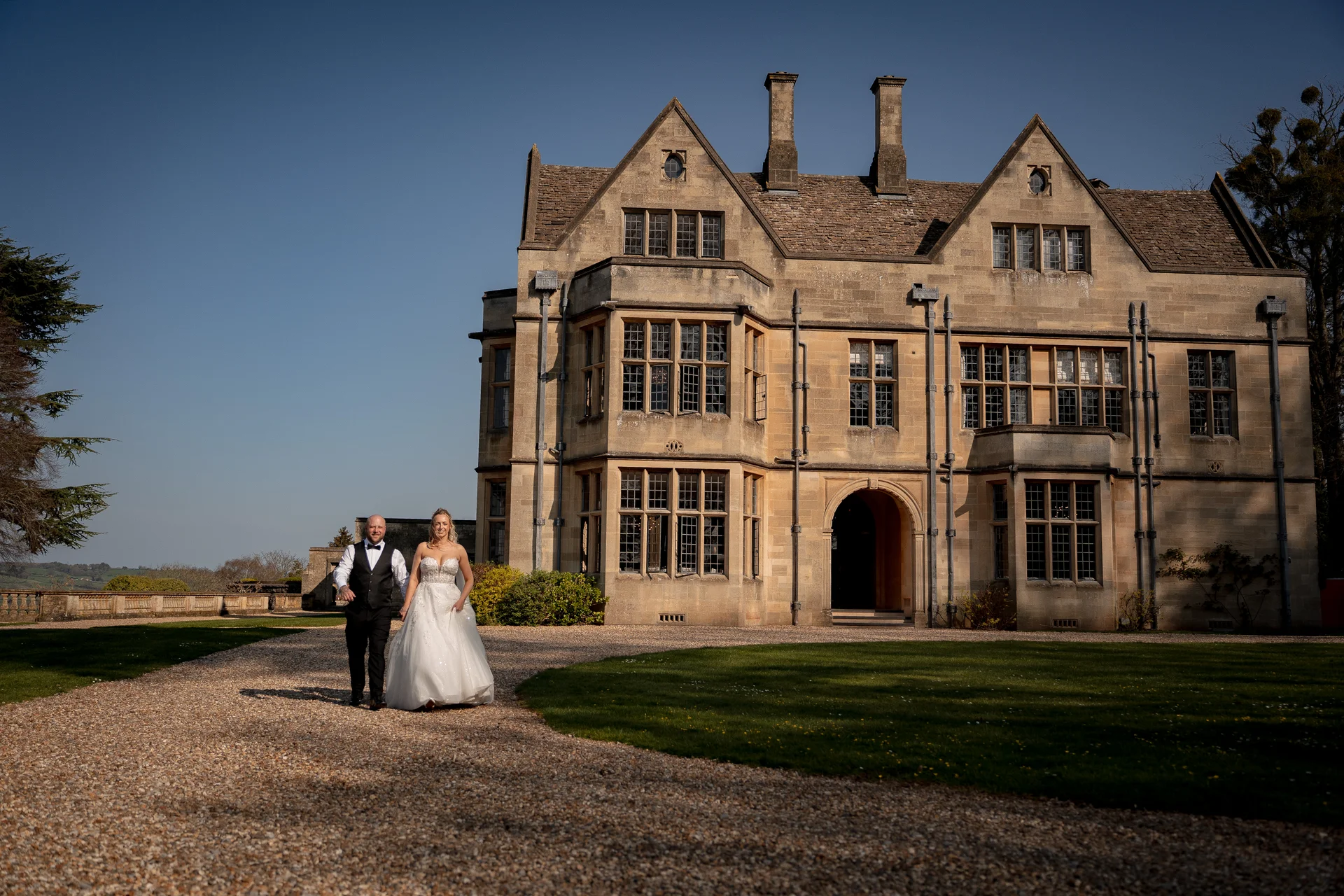 A newlywed couple walks hand in hand along a gravel driveway in front of a grand stone country manor house under a clear blue sky.