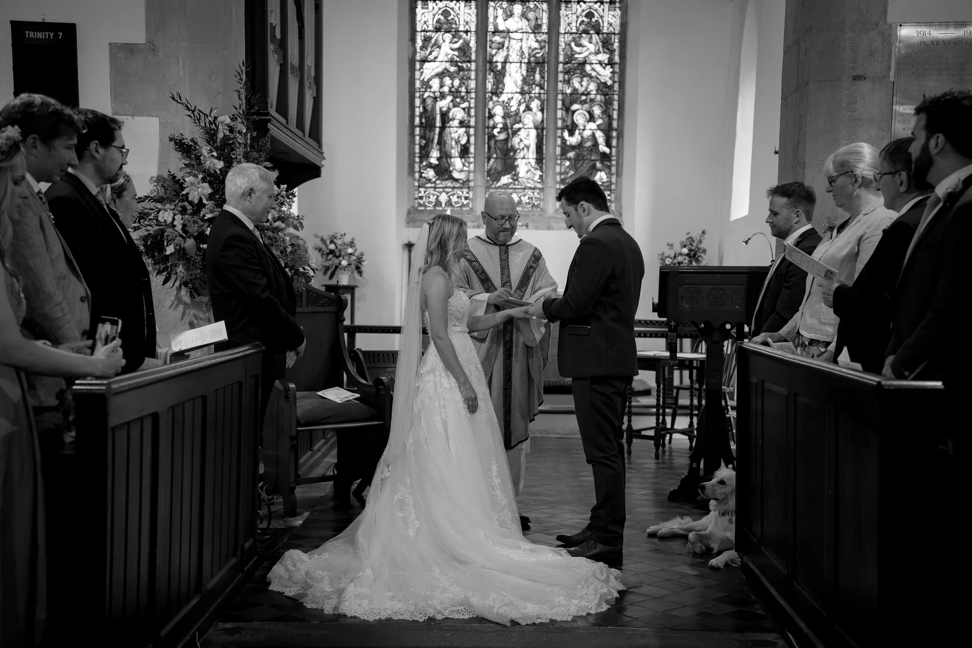 A bride and groom exchange rings at the altar of a historic church during a traditional ceremony, framed by stained glass windows and pews filled with guests.