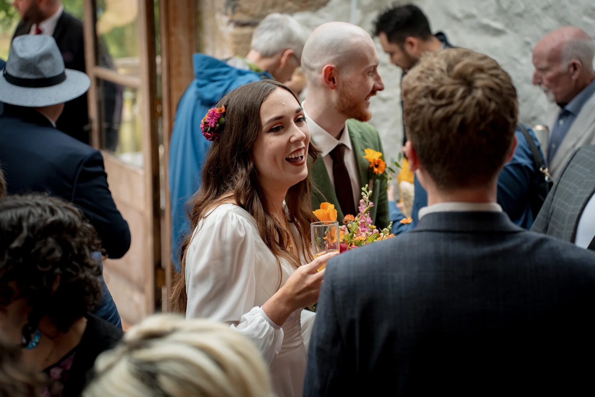 A smiling bride wearing a white dress with flowers in her hair laughs and holds a drink while mingling with wedding guests during a drinks reception.
