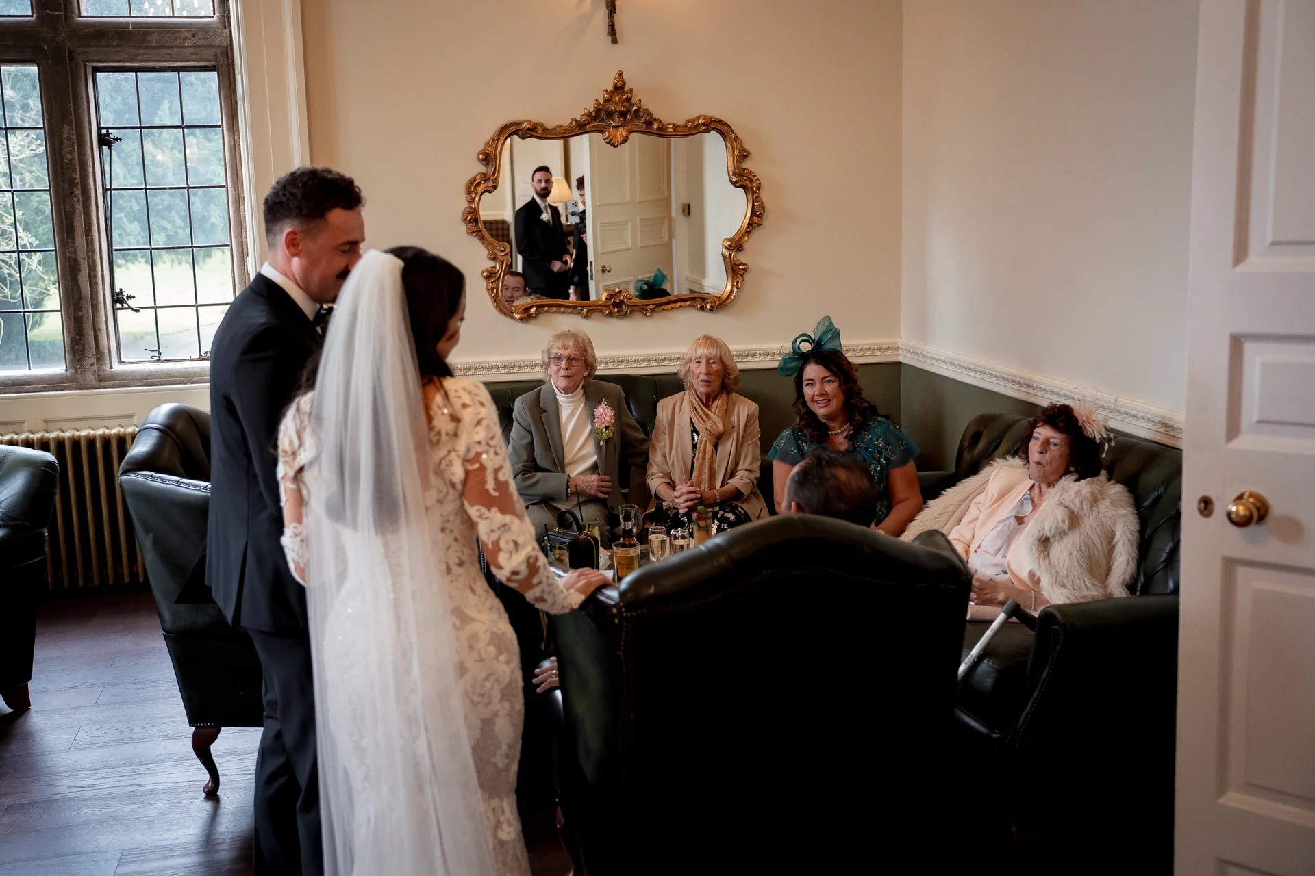 A bride in a lace gown with a veil stands with the groom in a classic suit, interacting with seated family members and guests in a light-filled country house sitting room.