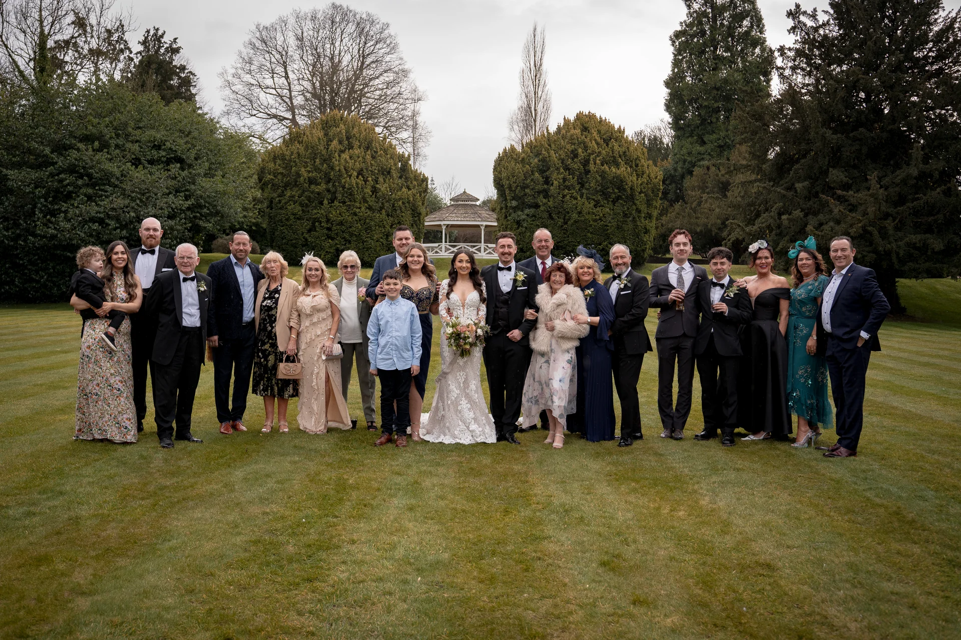 A large wedding party and family group pose formally on a pristine lawn in front of a distant gazebo under overcast skies.