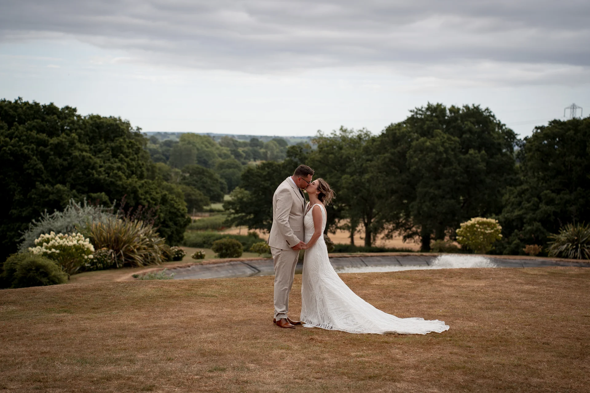 A newlywed couple shares a tender kiss outdoors on a grassy lawn overlooking a scenic landscape with rolling hills and a fountain under an overcast sky.