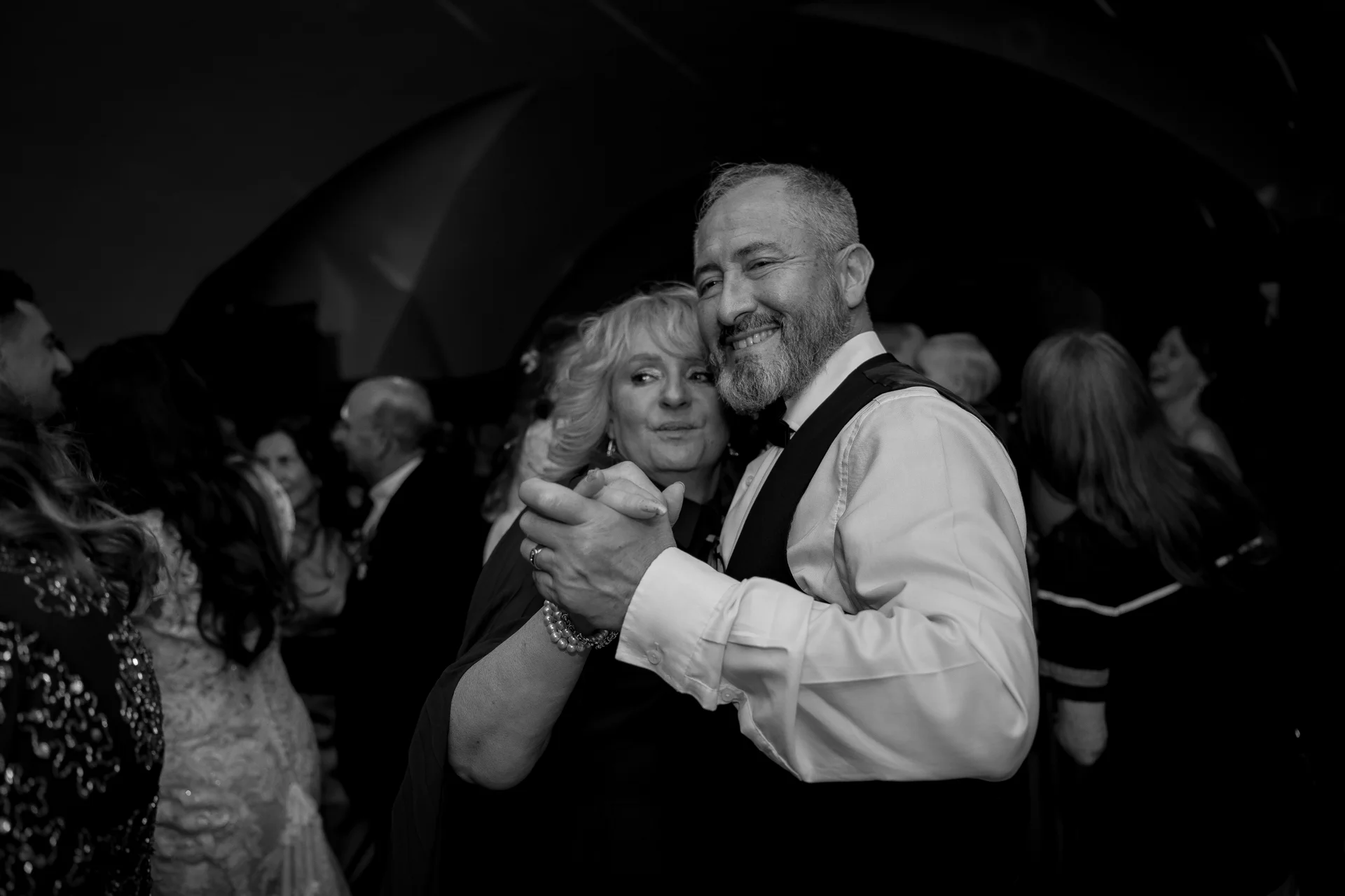 An older couple shares a joyful dance on a crowded reception dance floor, captured in a striking black and white photograph.