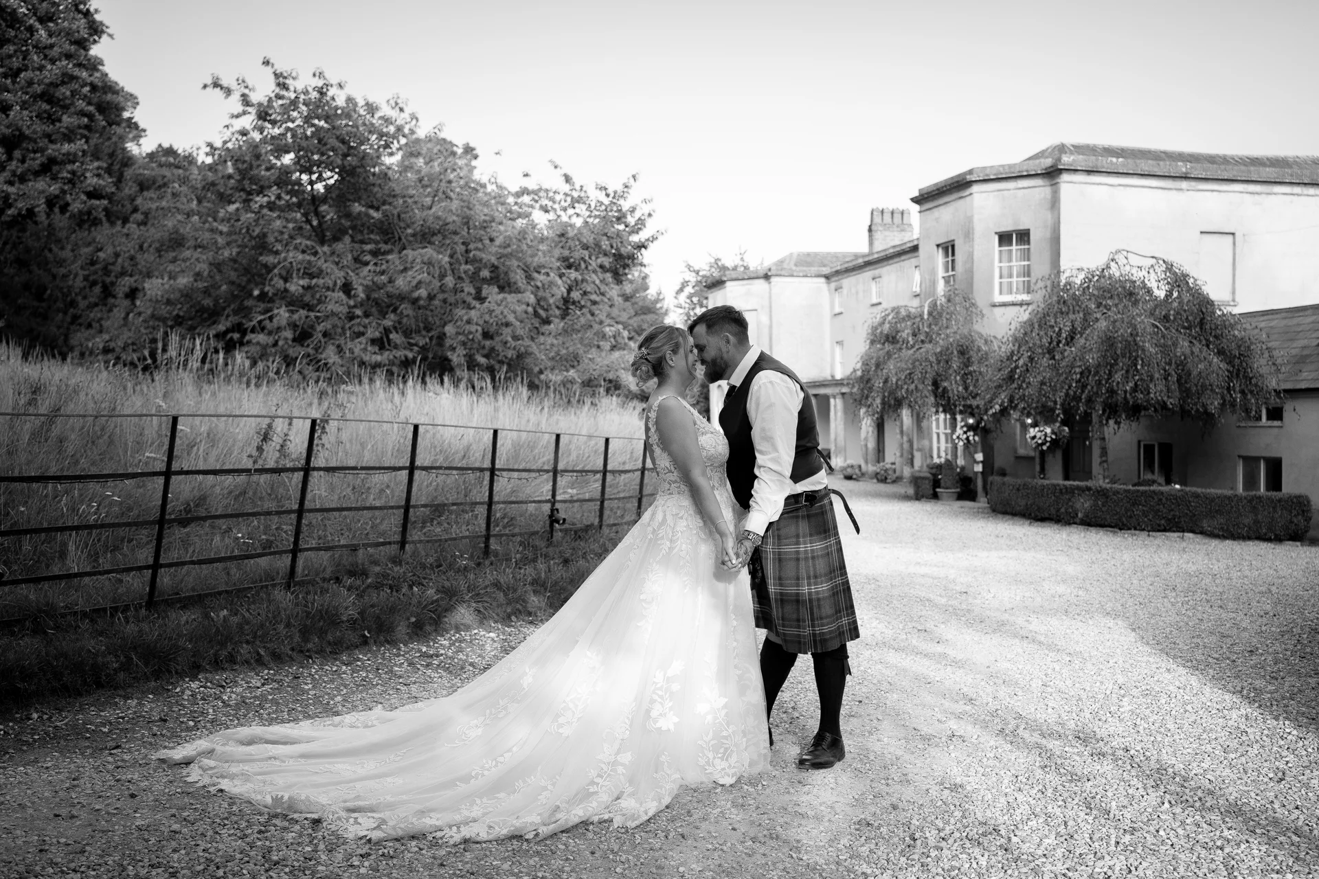 A newlywed couple share an intimate moment nose-to-nose outdoors on a gravel pathway in front of a grand country house, with the bride wearing a lace gown with a long train and the groom dressed in a traditional tartan kilt.