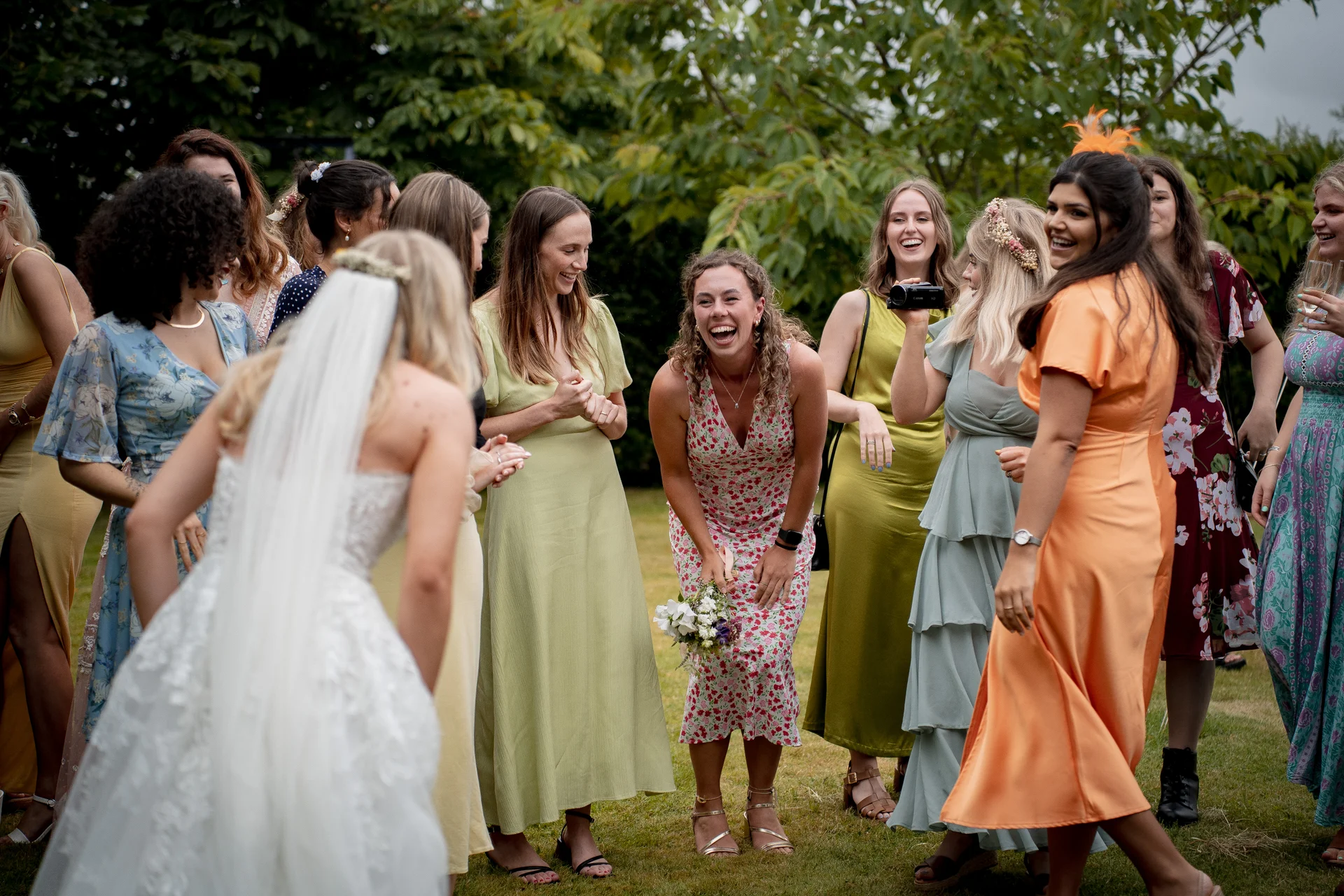 The bride in a white lace gown and veil interacts with a joyful group of female guests and bridesmaids laughing heartily outdoors in a lush garden setting.