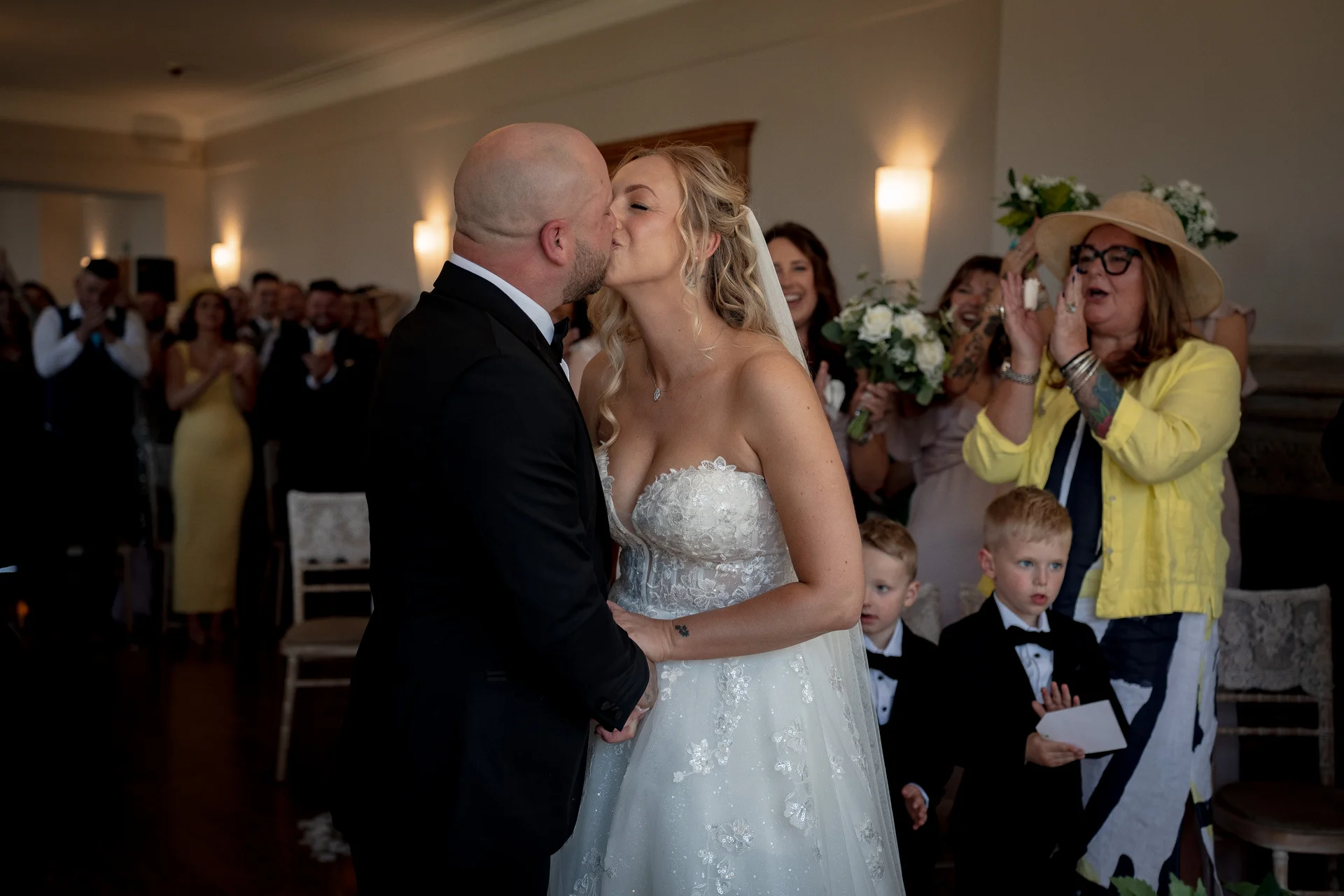 A newly married couple share their first kiss at the altar while guests applaud joyfully in the background.