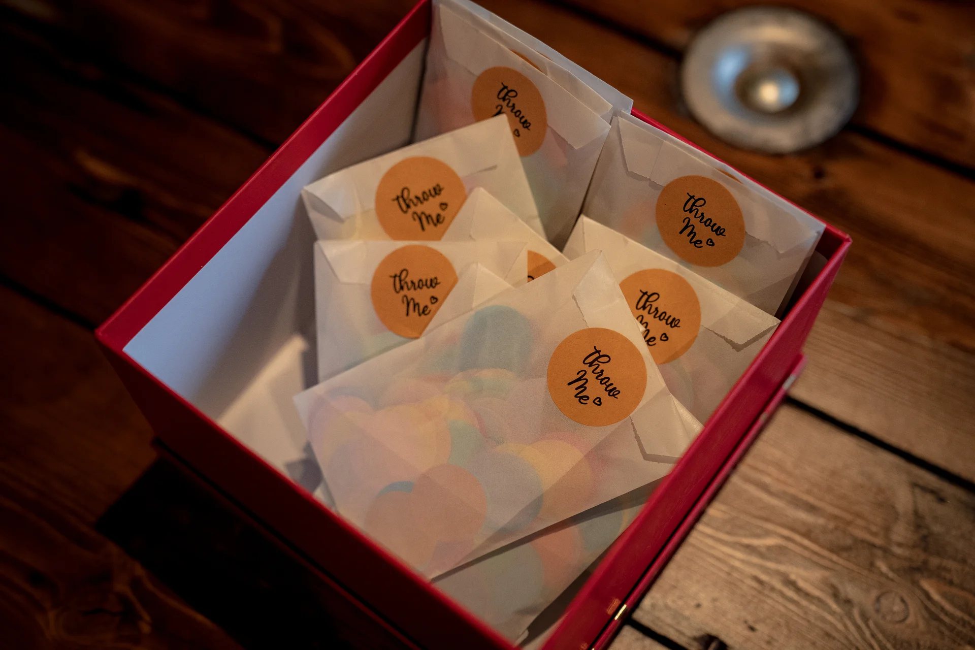 A red storage box filled with translucent paper confetti envelopes featuring round throw me stickers sits neatly on a rustic wooden table.