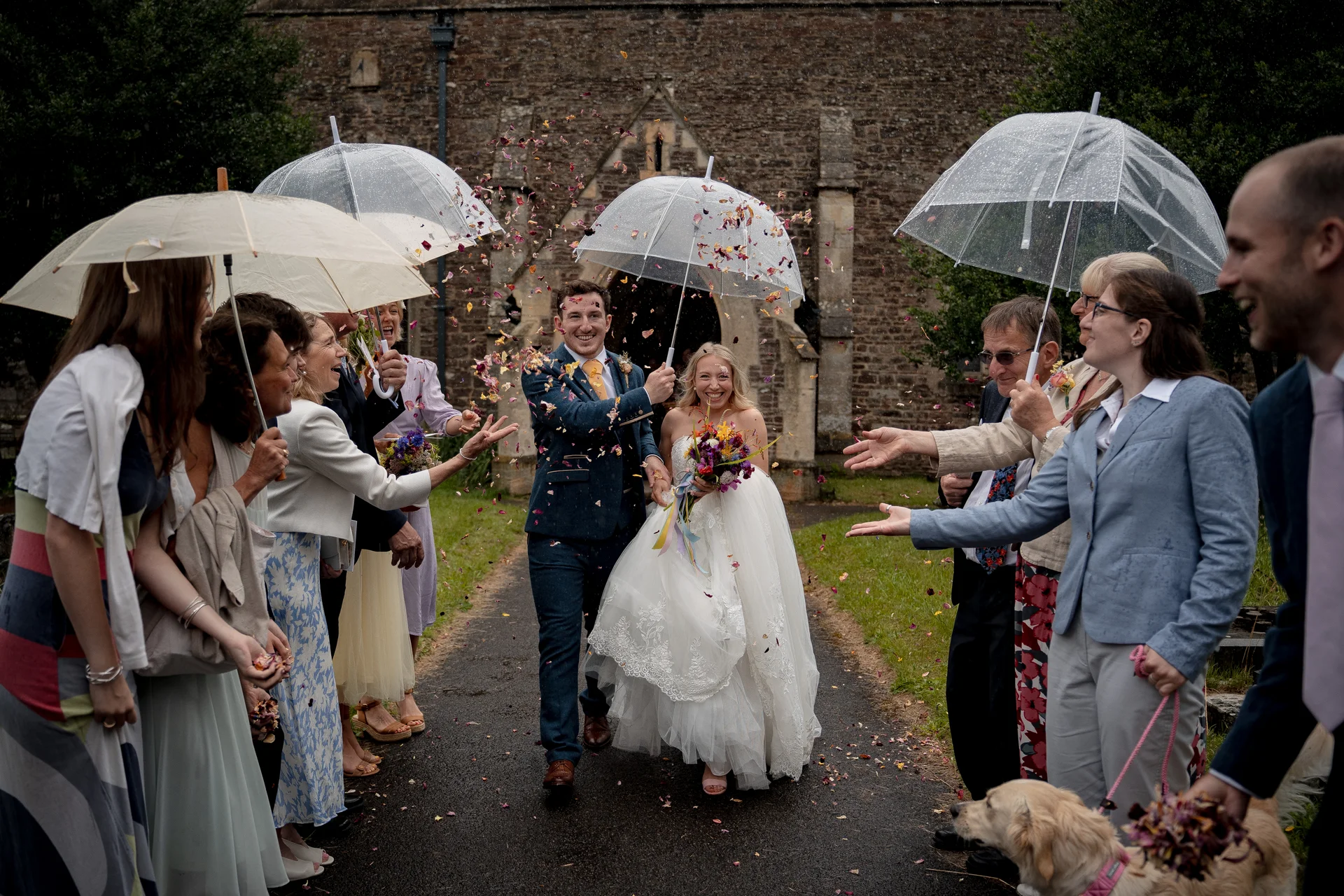 A newlywed couple laugh as guests shower them with petal confetti outside a stone church while holding clear umbrellas on a rainy day.