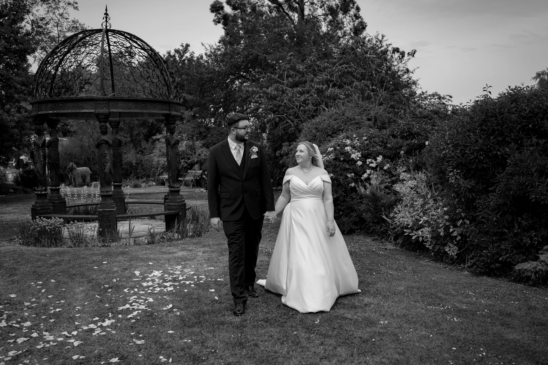 A newlywed couple holds hands and shares a loving glance while walking across a garden lawn near an ornate metal gazebo.