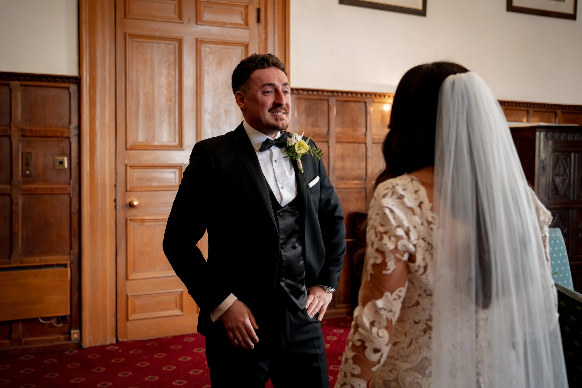 An emotional groom becomes tearful with joy during a first look moment with his bride inside a wood-panelled room.