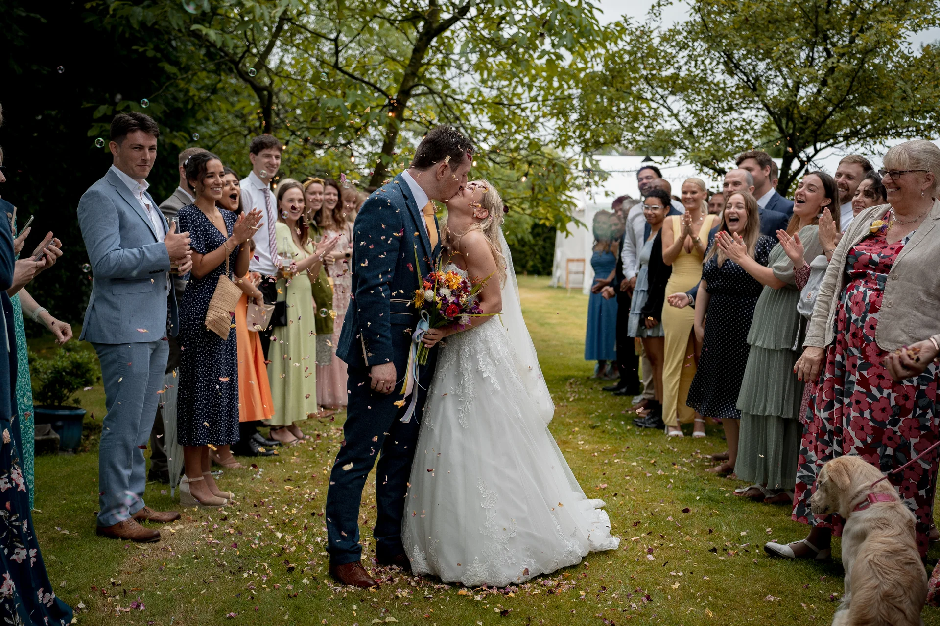 Newlywed couple shares a romantic kiss as cheerful wedding guests applaud, showering them with colourful petal confetti outdoors in a lush garden setting.