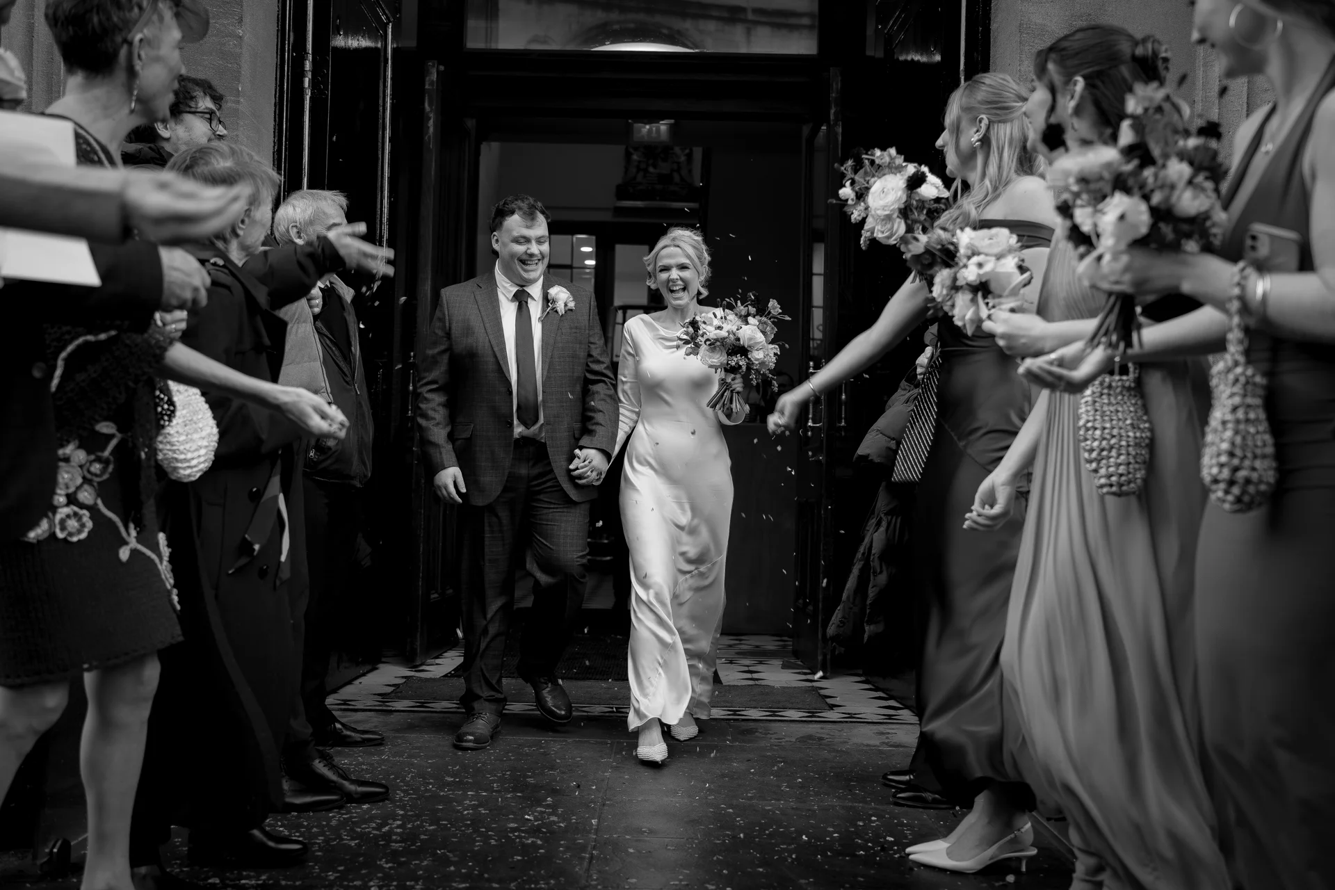 A joyous newlywed couple exits a grand doorway hand in hand amidst a shower of confetti, surrounded by cheering guests and bridesmaids holding bouquets in a monochrome documentary shot.