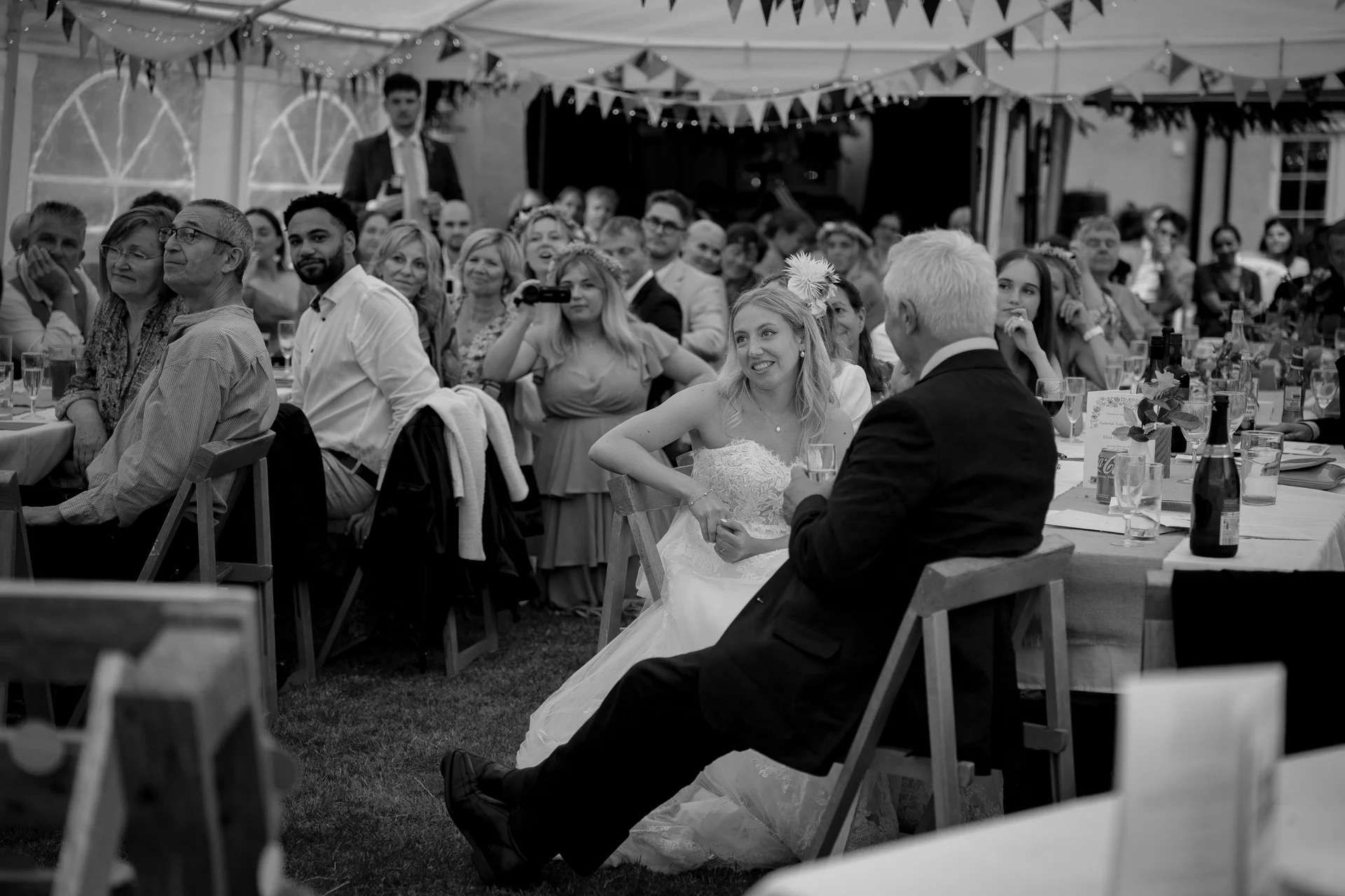 A bride in a lace wedding dress and a guest share a joyful moment during speeches inside a marquee adorned with bunting.