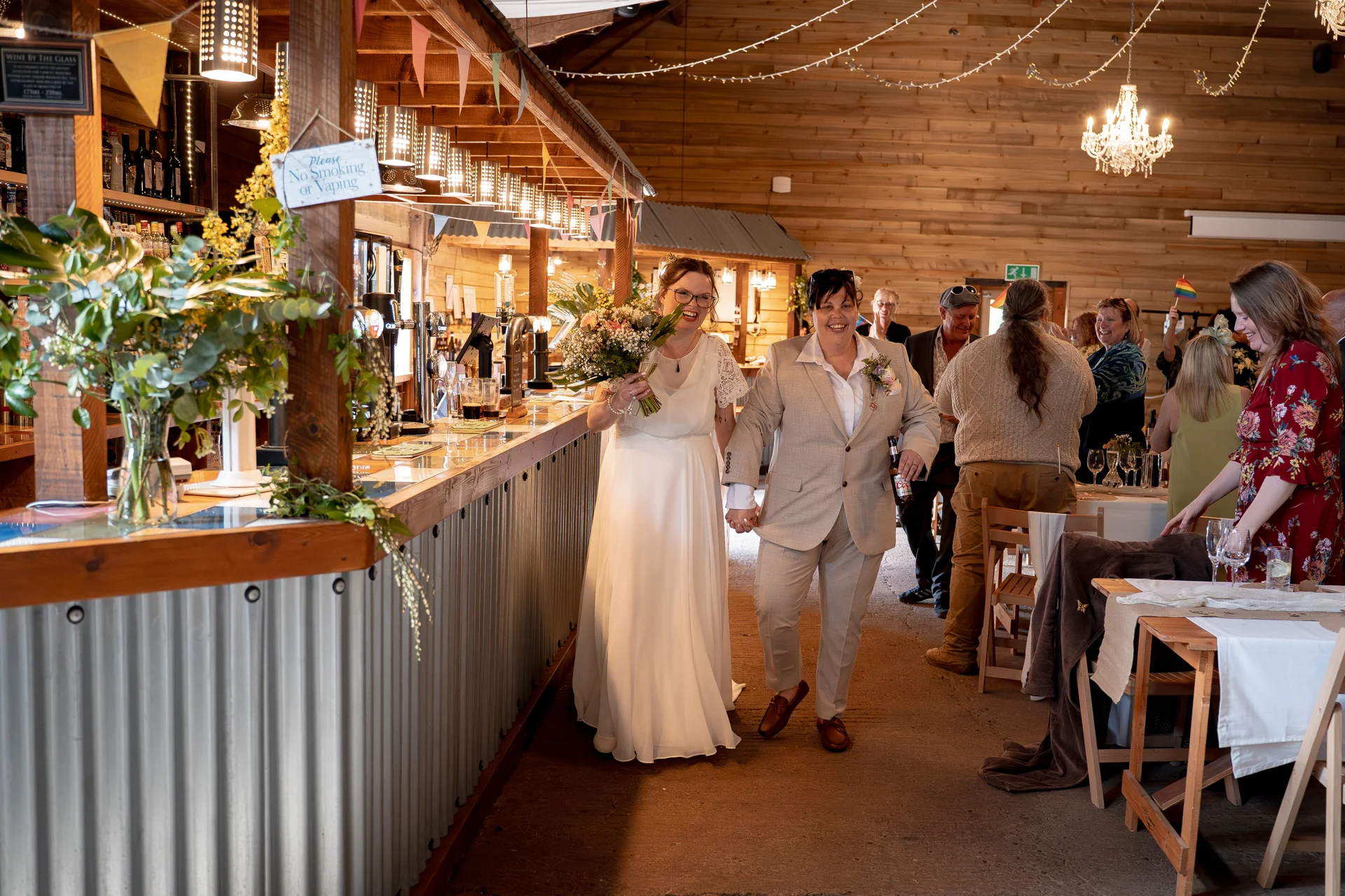 A newlywed same-sex couple joyfully walks hand in hand through a rustic wooden barn venue past a bar while guests look on and smile.