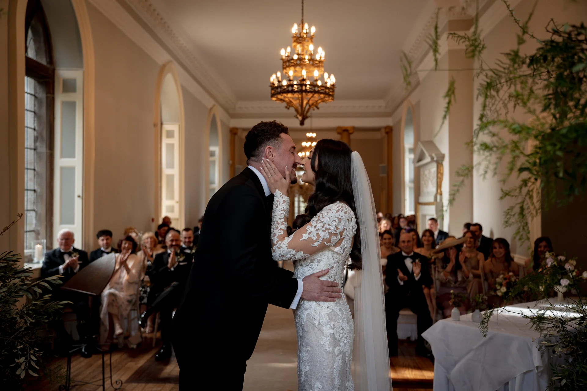 Newlywed bride and groom share a romantic first kiss inside an elegant manor house ceremony room while guests applaud in the background.