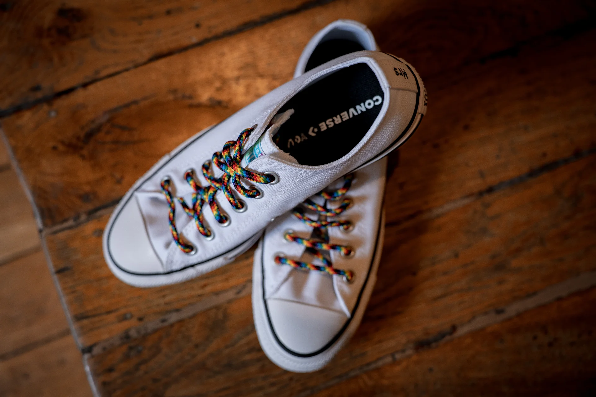 A pair of white Converse bridal trainers featuring vibrant rainbow laces rest on a rustic wooden floor.