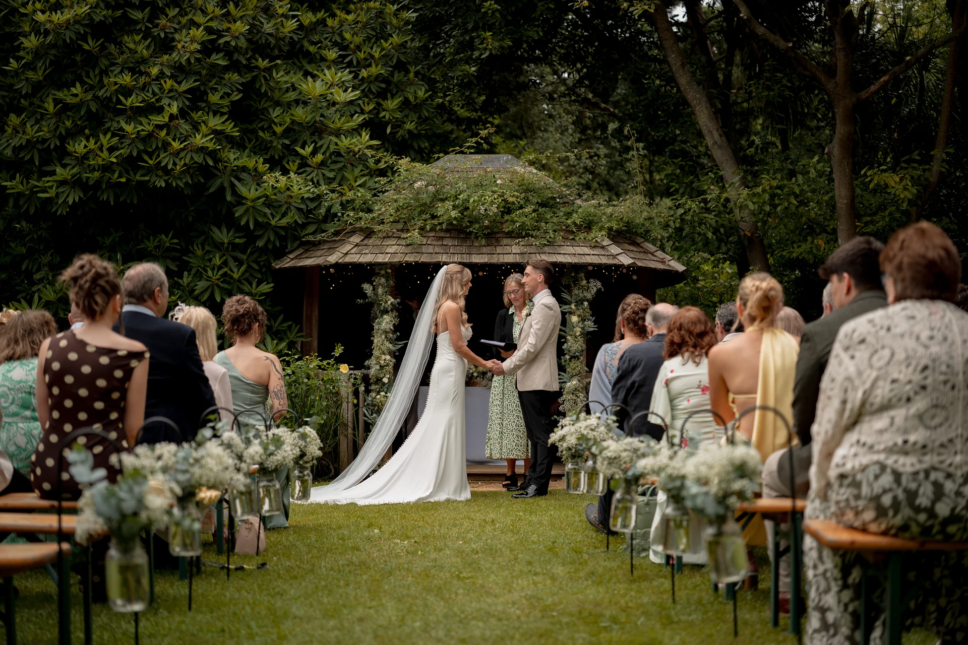 A bride and groom exchange vows in front of a rustic wooden gazebo decorated with foliage, surrounded by seated guests during an outdoor summer wedding ceremony.