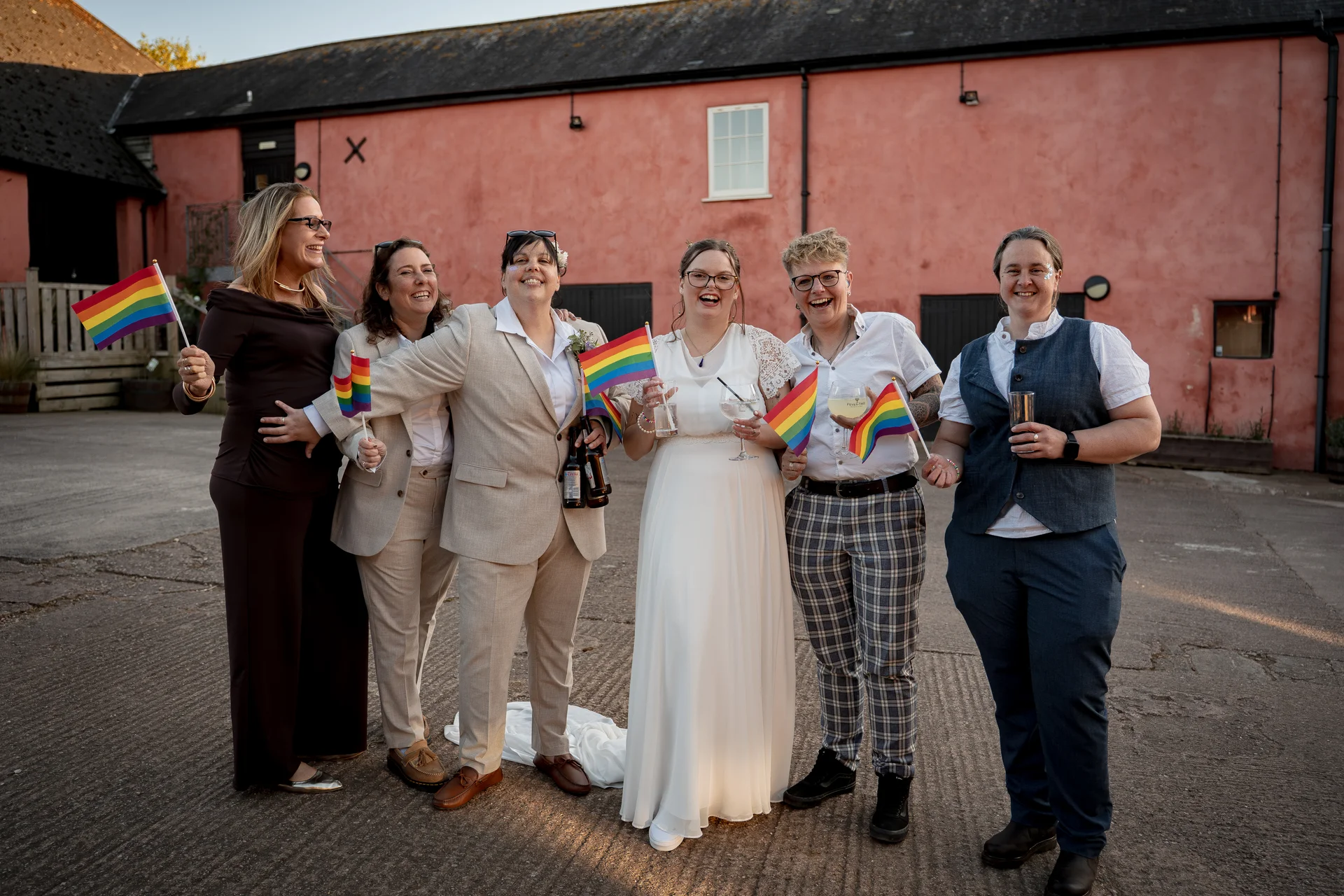 A smiling bride in a white dress and her wedding party hold small rainbow flags and drinks while posing together in a rustic brick courtyard.