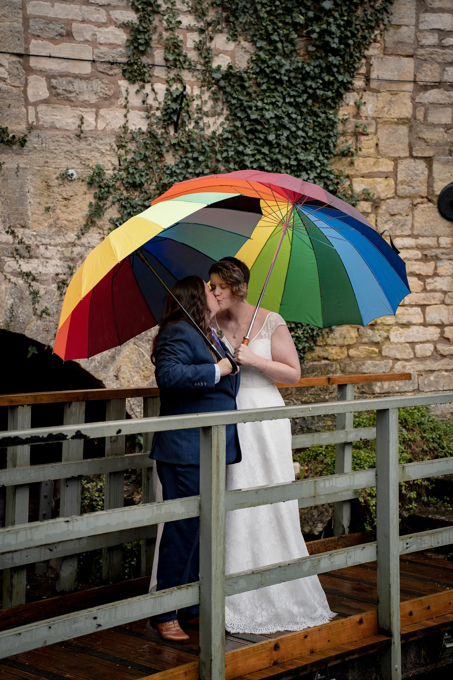 Two brides share a tender kiss beneath a colourful rainbow umbrella while standing on a wooden footbridge against an ivy-clad stone wall.