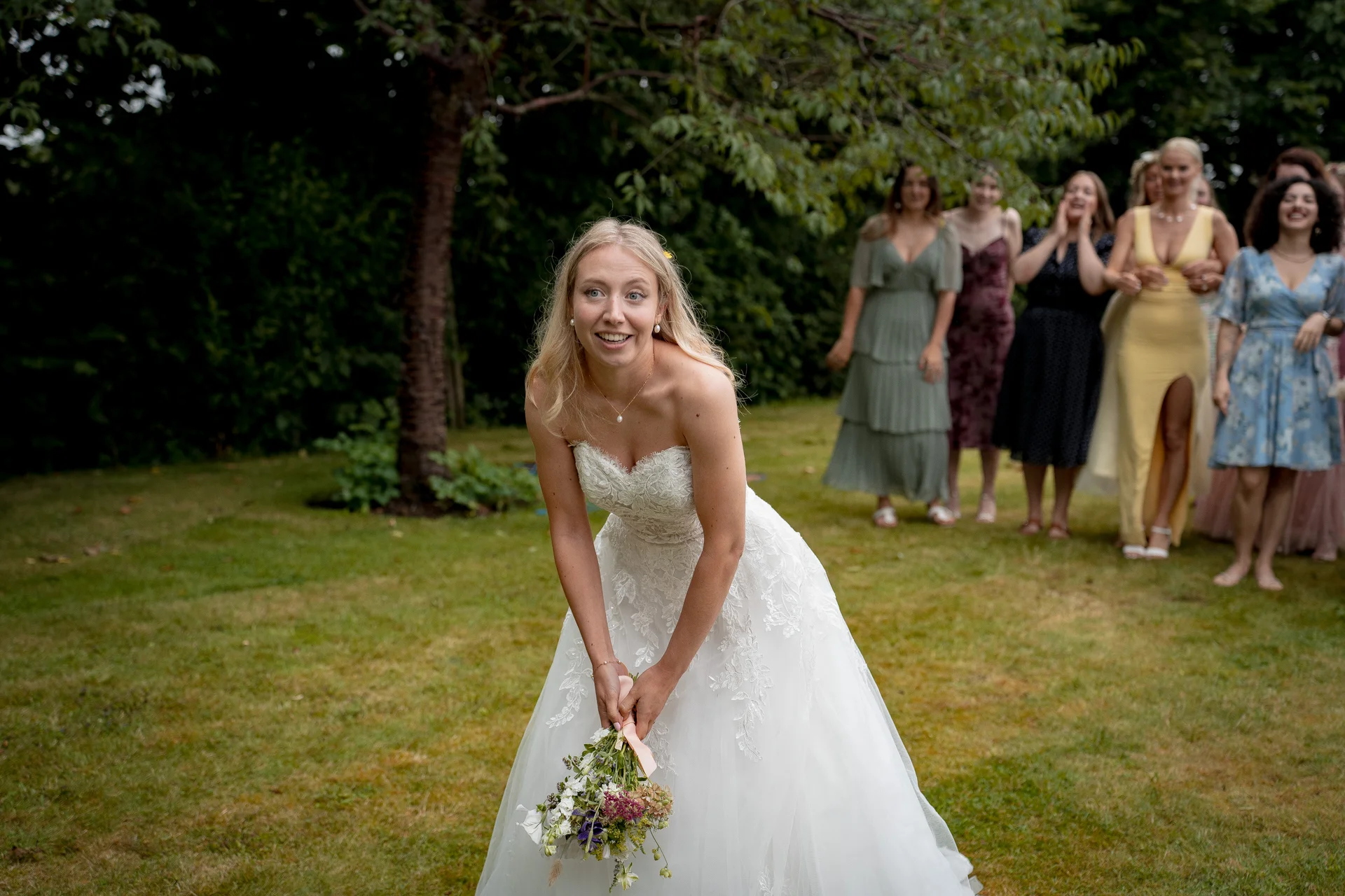 A joyful bride leans forward on a grassy lawn while preparing to toss her bouquet, with an excited group of bridesmaids watching in the background.