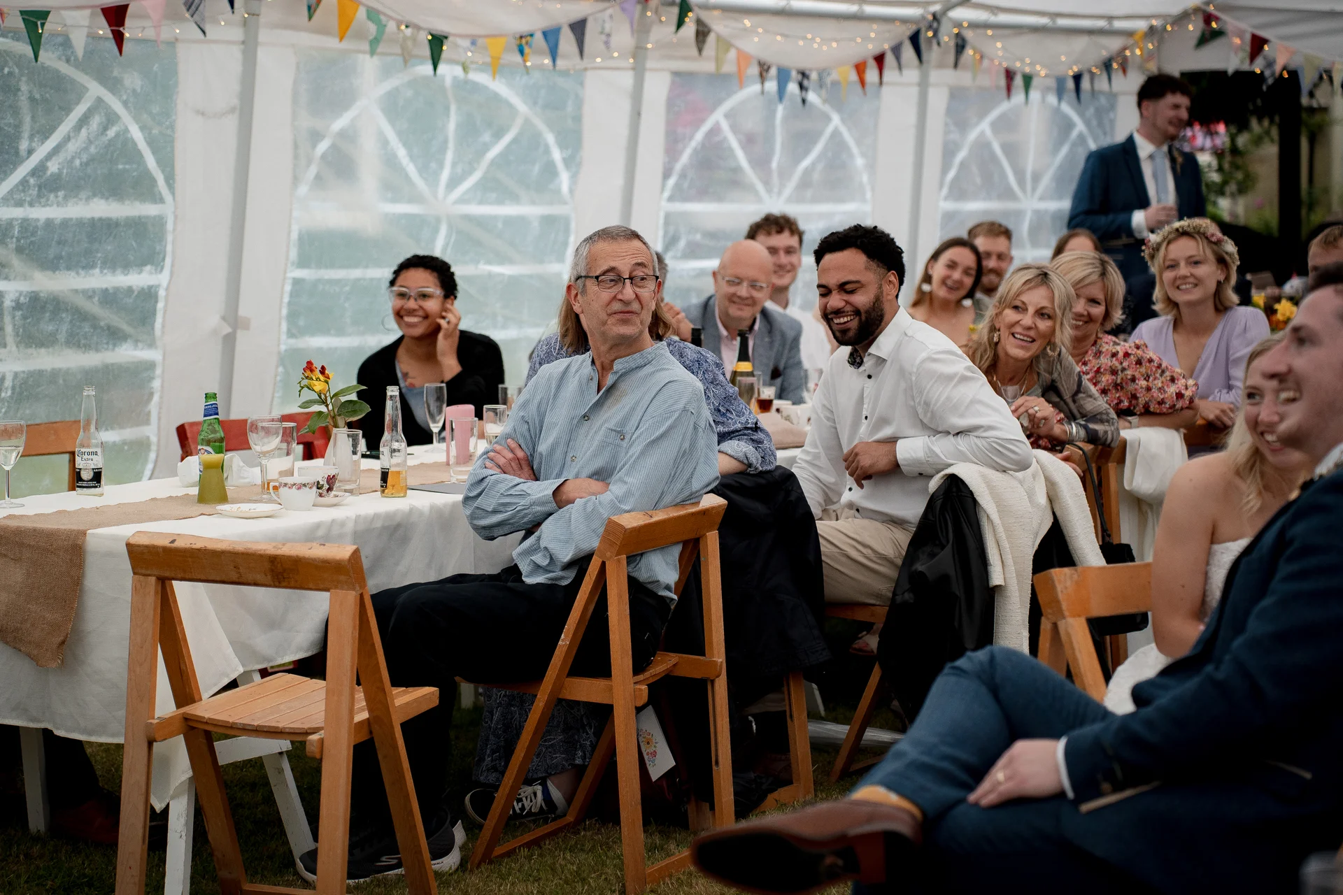 A joyful group of wedding guests seated inside a decorated marquee with bunting and fairy lights, laughing and watching the wedding speeches.