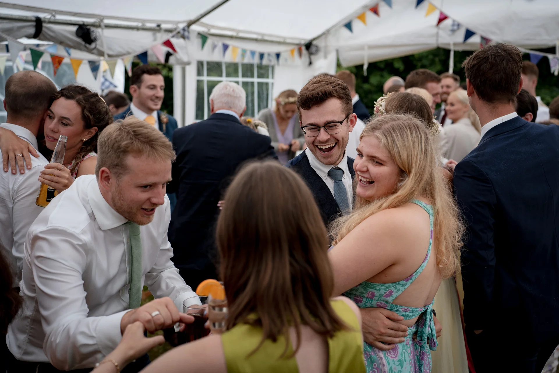 Wedding guests mingle and laugh joyfully inside a brightly lit marquee decorated with colourful bunting during a lively drinks reception.