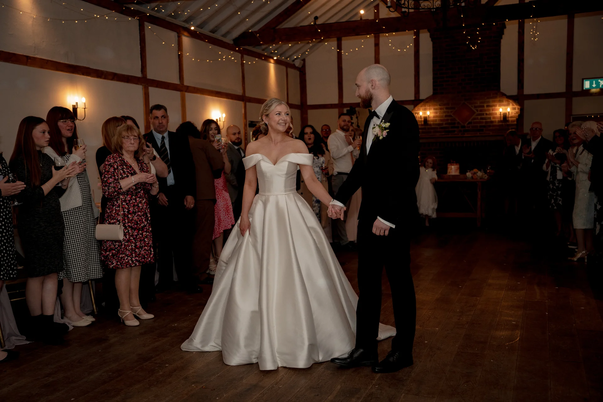 The newlywed couple share their first dance in a rustic oak-framed barn decorated with fairy lights, surrounded by watching and applauding guests.