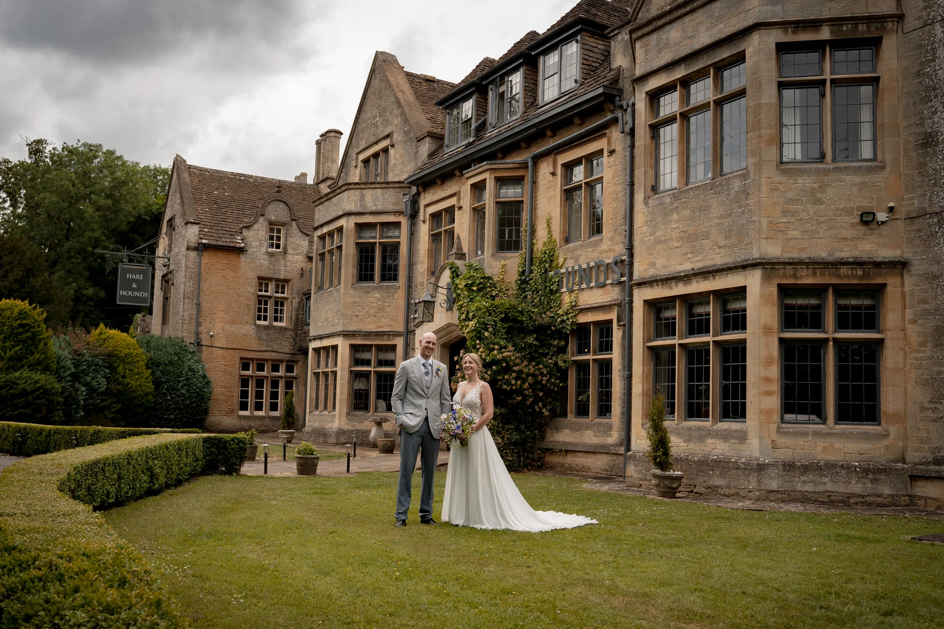 A newlywed couple stands on the lawn in front of a historic stone manor house featuring large mullioned windows and manicured gardens.