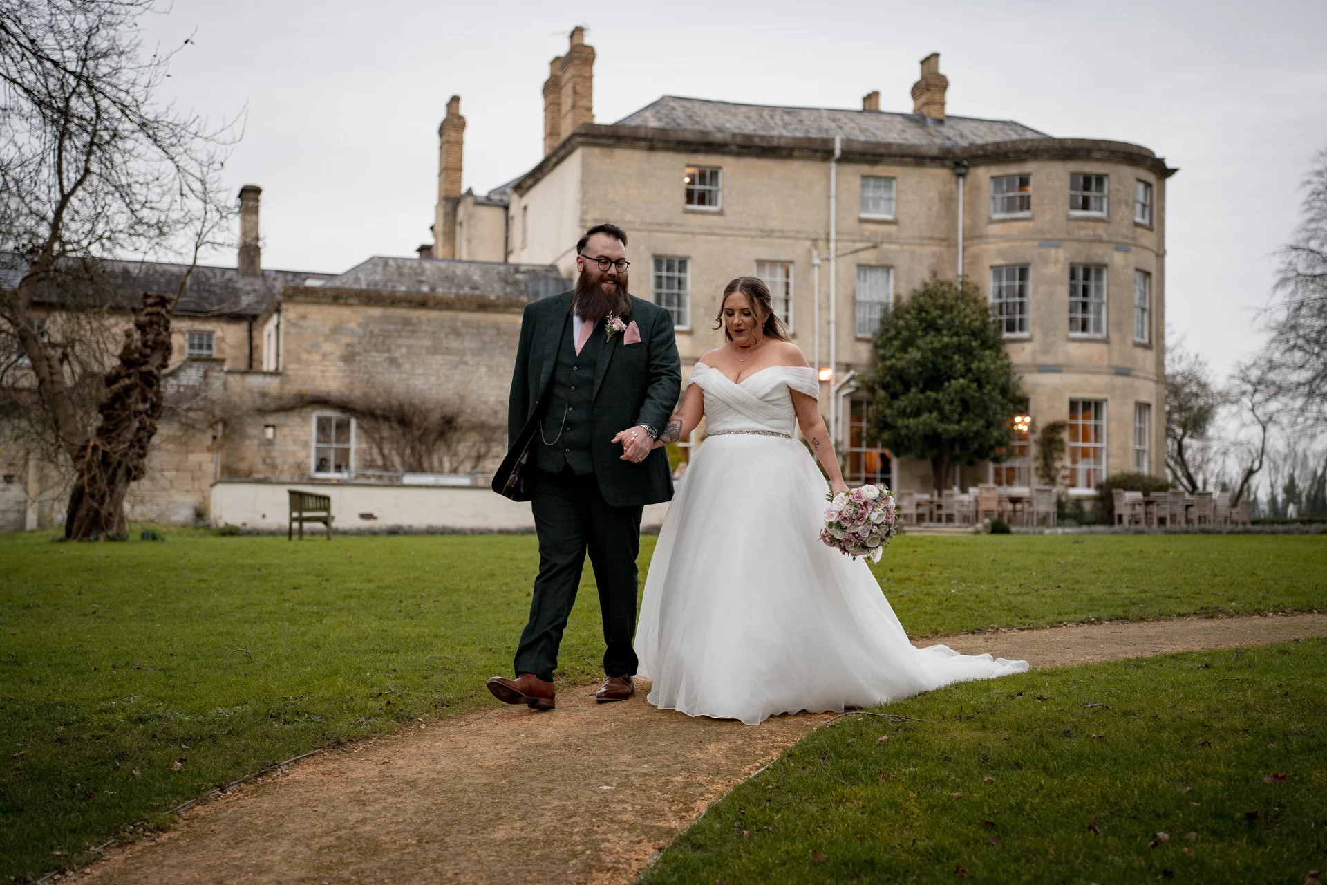 A newlywed couple walks hand in hand along a gravel path across a lush green lawn in front of a grand stone country house.