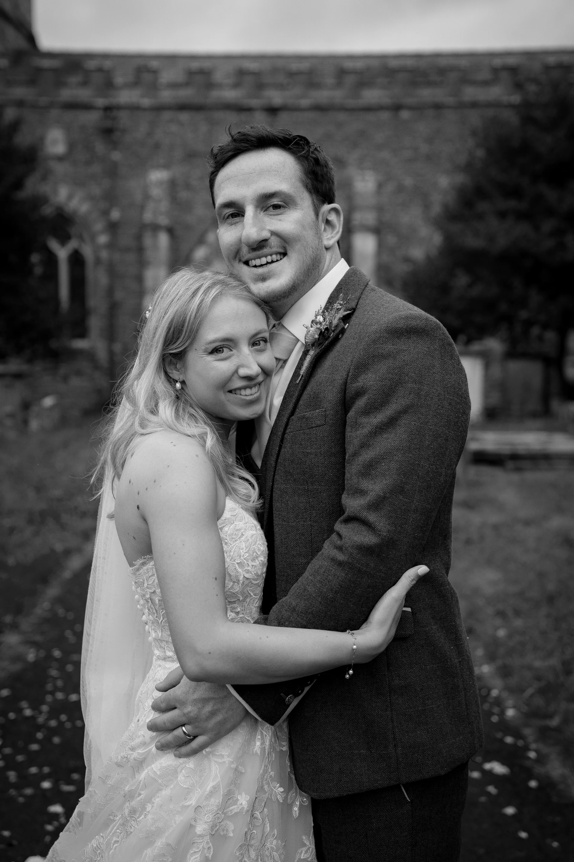 A smiling bride and groom embrace closely outside a historic stone church on their wedding day.