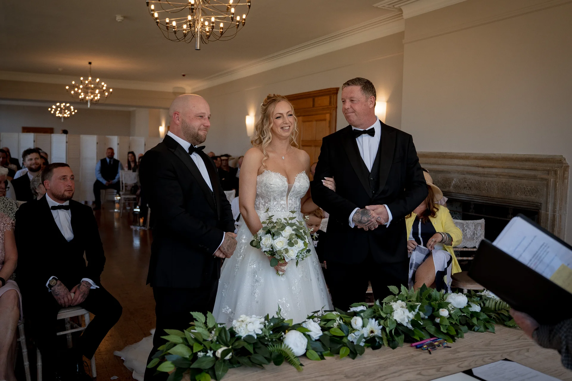 A smiling bride holding a white rose bouquet stands at the altar between two groomsmen wearing black tuxedos during an indoor civil ceremony.