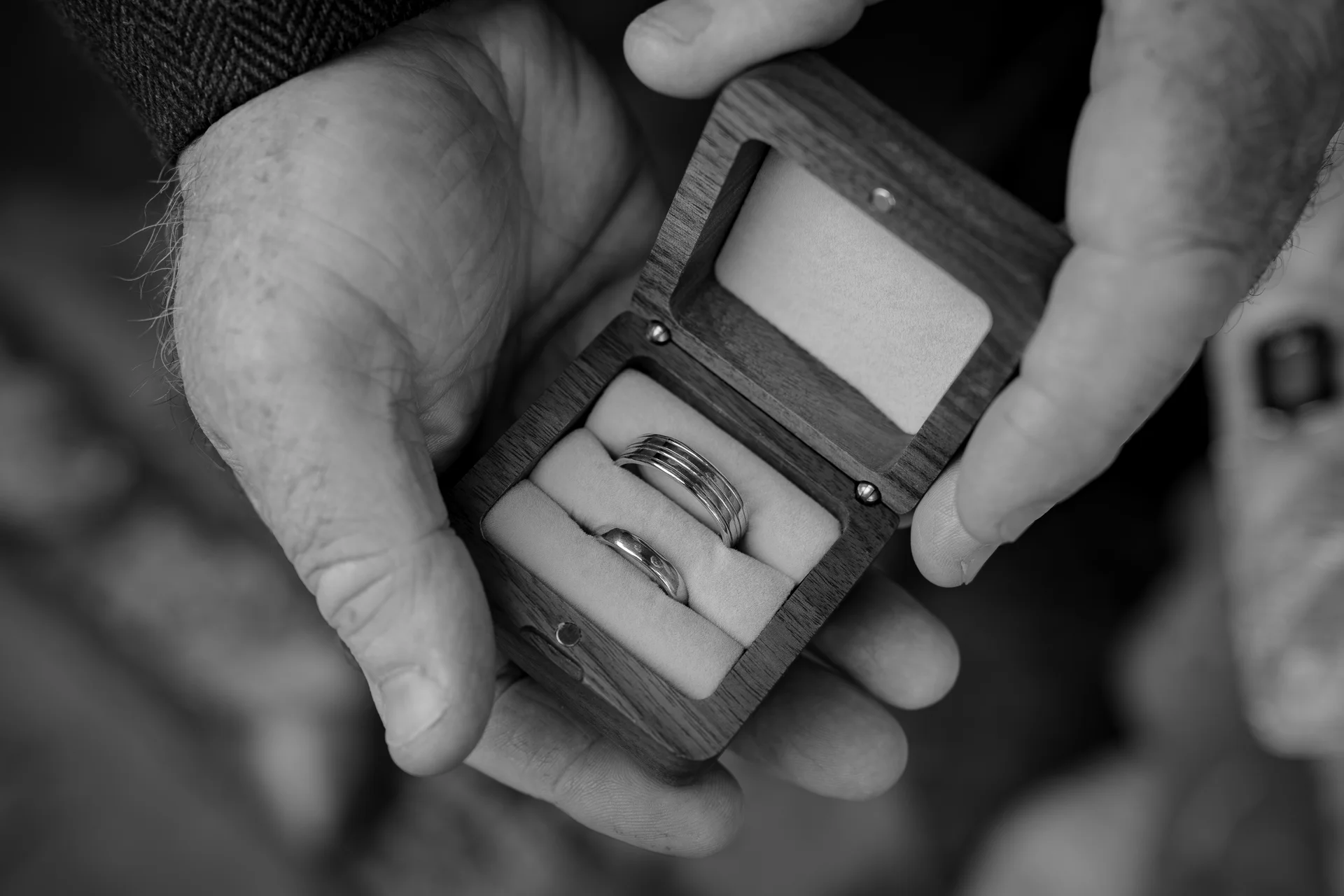 Close-up black and white photograph of hands gently holding an open wooden ring box containing two distinct wedding bands.