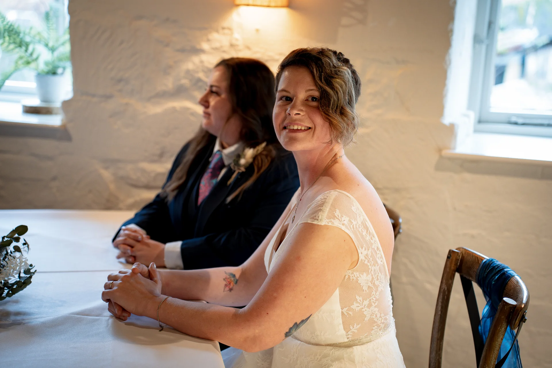 A smiling bride in a lace wedding dress sits at the top table next to her wife, who wears a smart suit, inside a rustic venue with white stone walls.