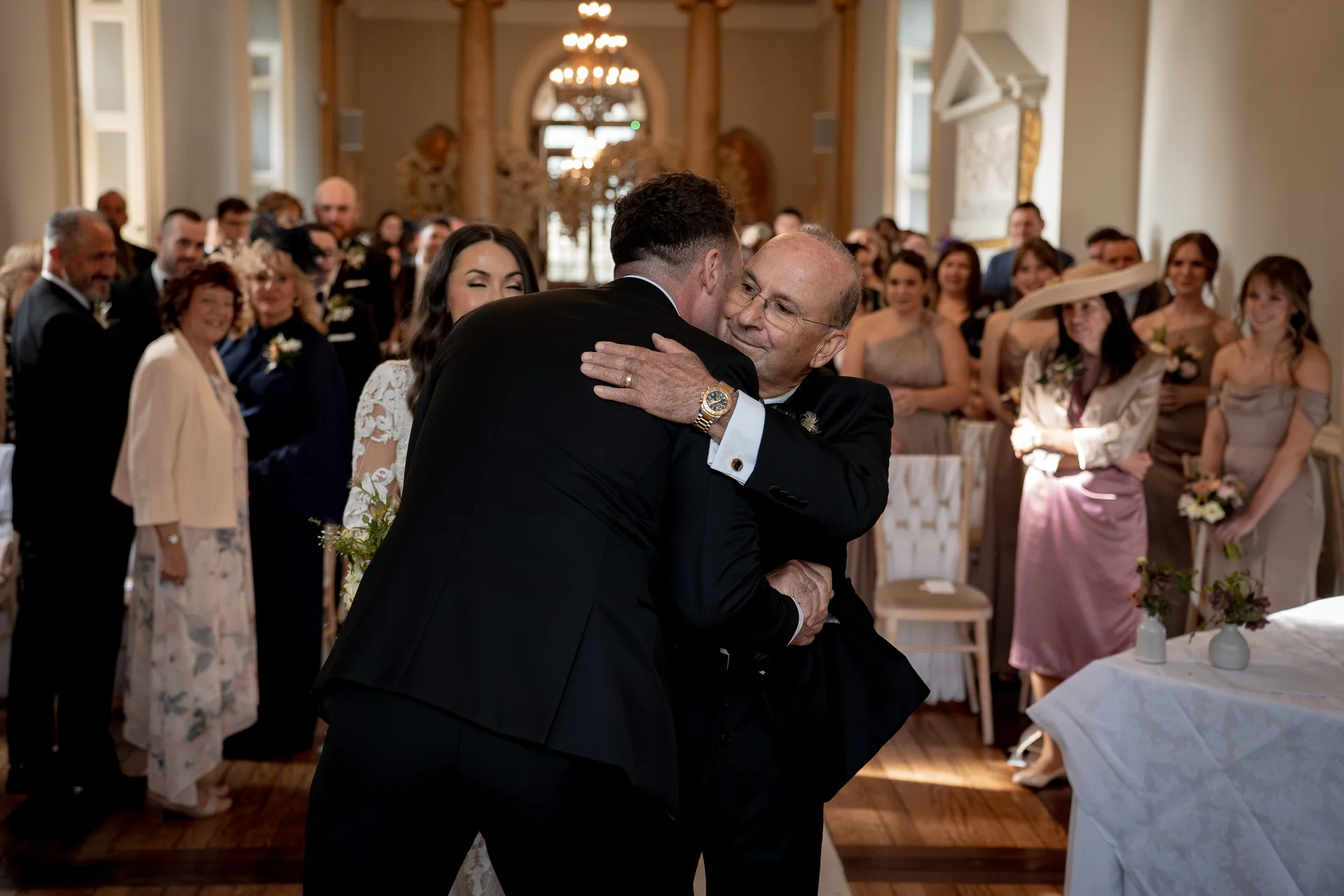 The groom warmly hugs the father of the bride at the front of a light-filled ceremony room while guests watch from their seats.