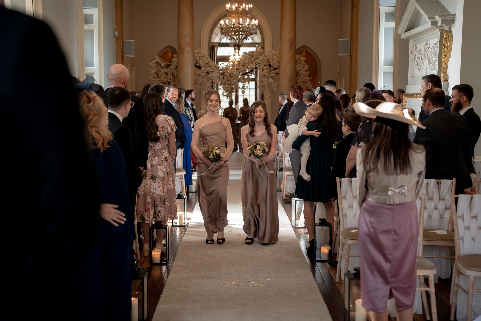 Two bridesmaids in champagne satin one-shoulder dresses walk down the aisle carrying bouquets during a country house wedding ceremony.