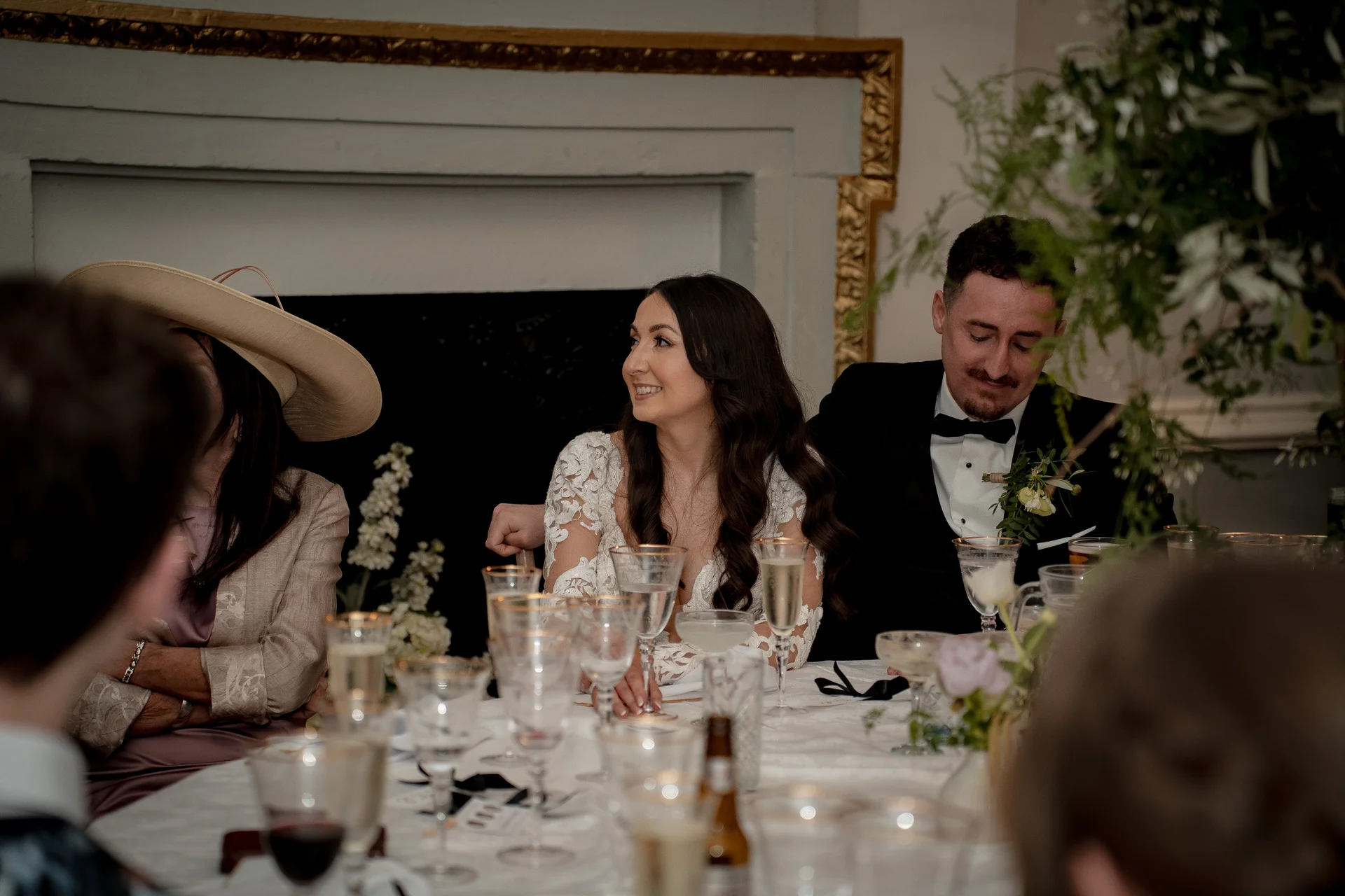 The bride and groom seated at the top table during wedding speeches, laughing together while guests enjoy the reception.