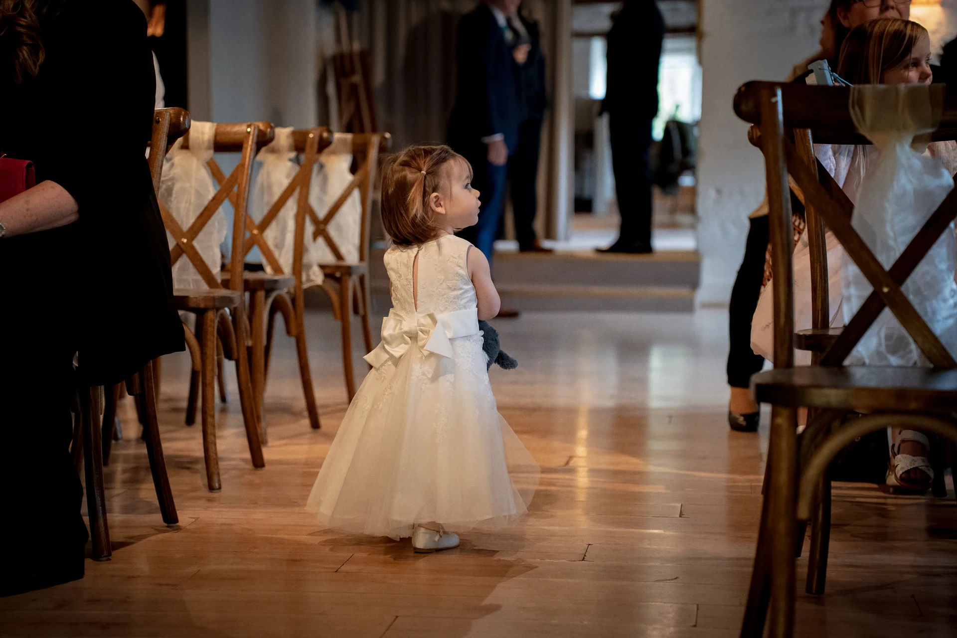 A young flower girl in a white dress with a bow stands in the aisle of a venue with wooden flooring, watching the ceremony.