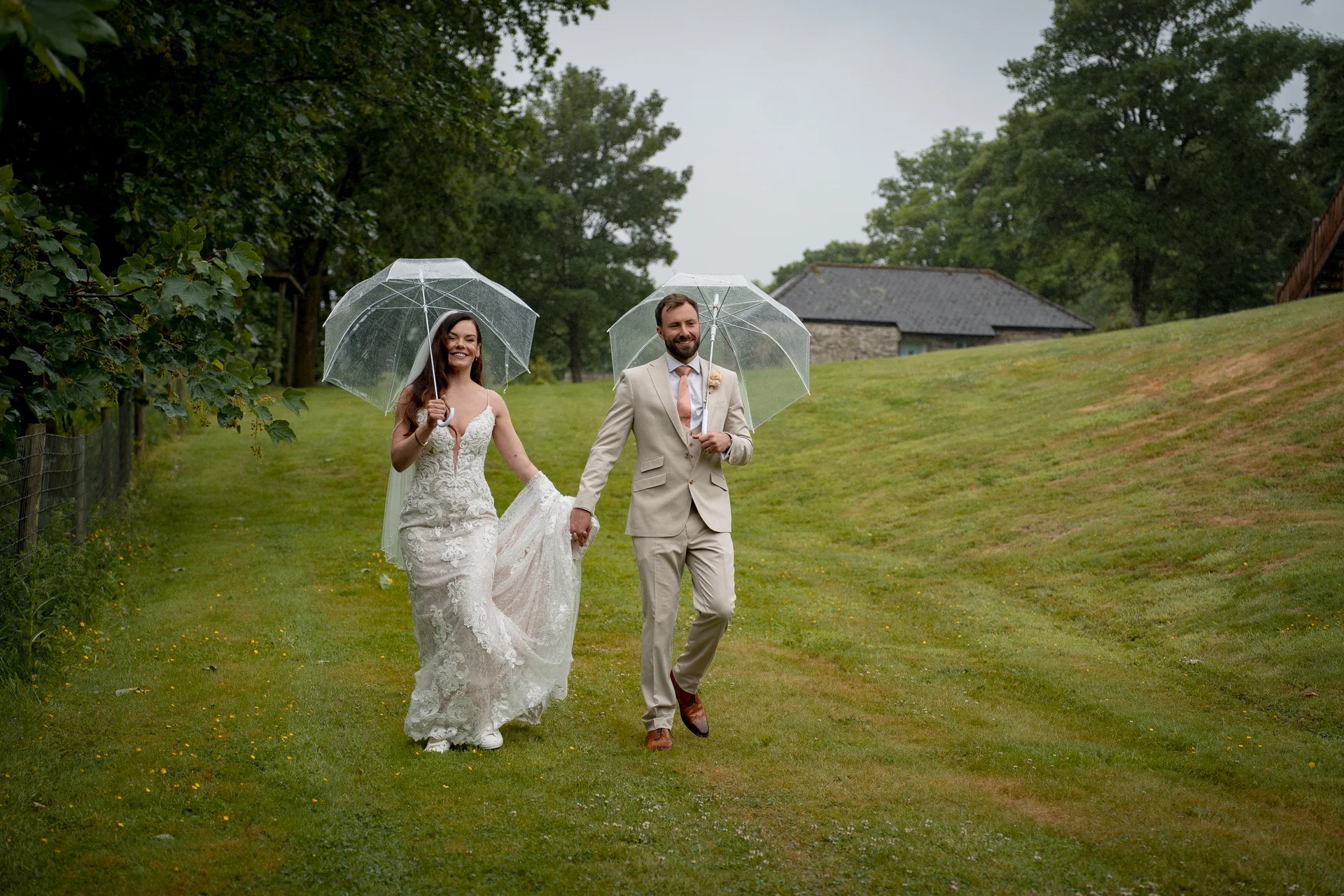 A smiling bride and groom walk hand in hand across a grassy hill under clear umbrellas on a rainy wedding day.