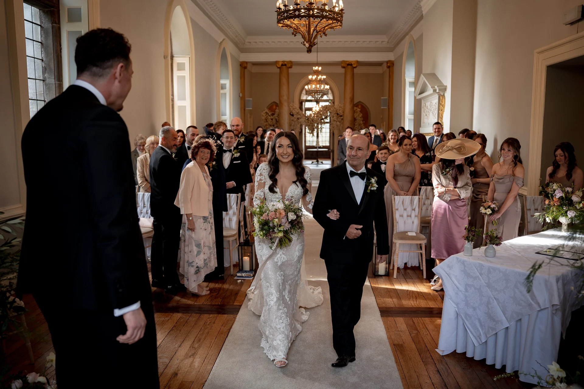 A smiling bride walks down the aisle on the arm of her father, towards the waiting groom in a grand hall.