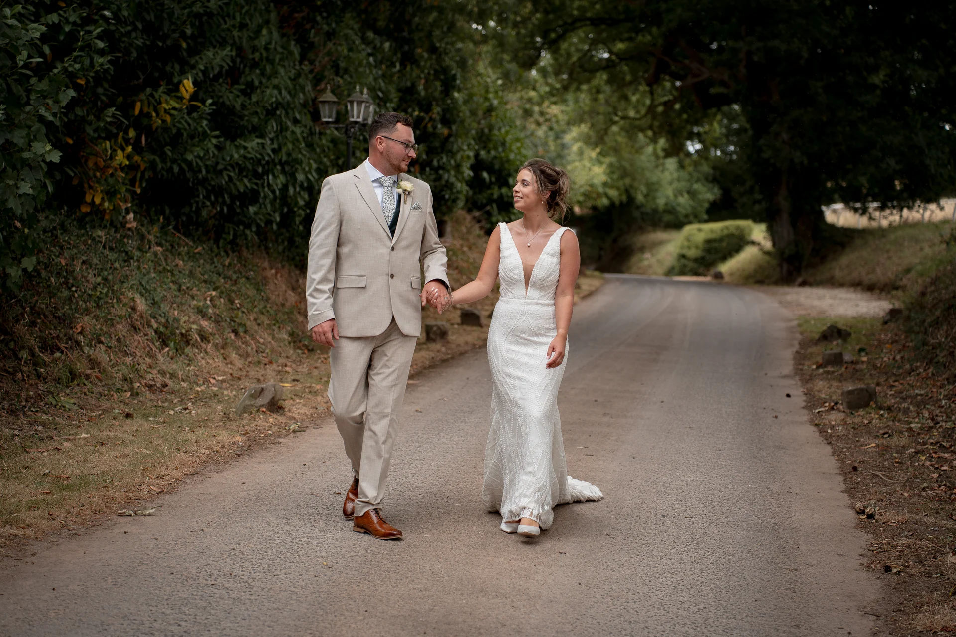 A bride and groom walk hand in hand down a quiet country lane, sharing a loving glance amidst lush greenery and trees.