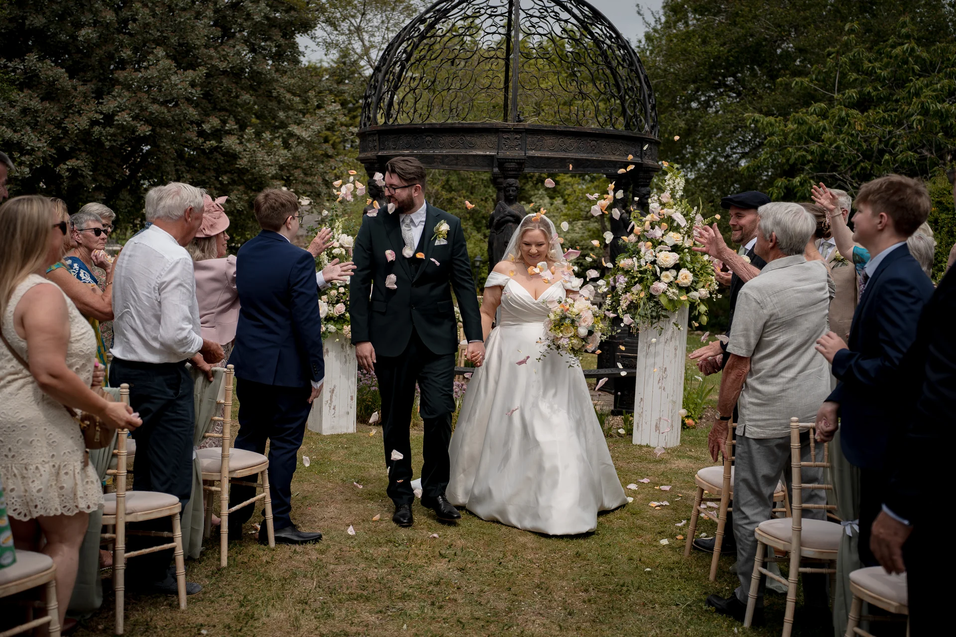 Newlyweds walk hand in hand down the aisle as guests happily throw confetti during an outdoor garden ceremony near a black metal bandstand.