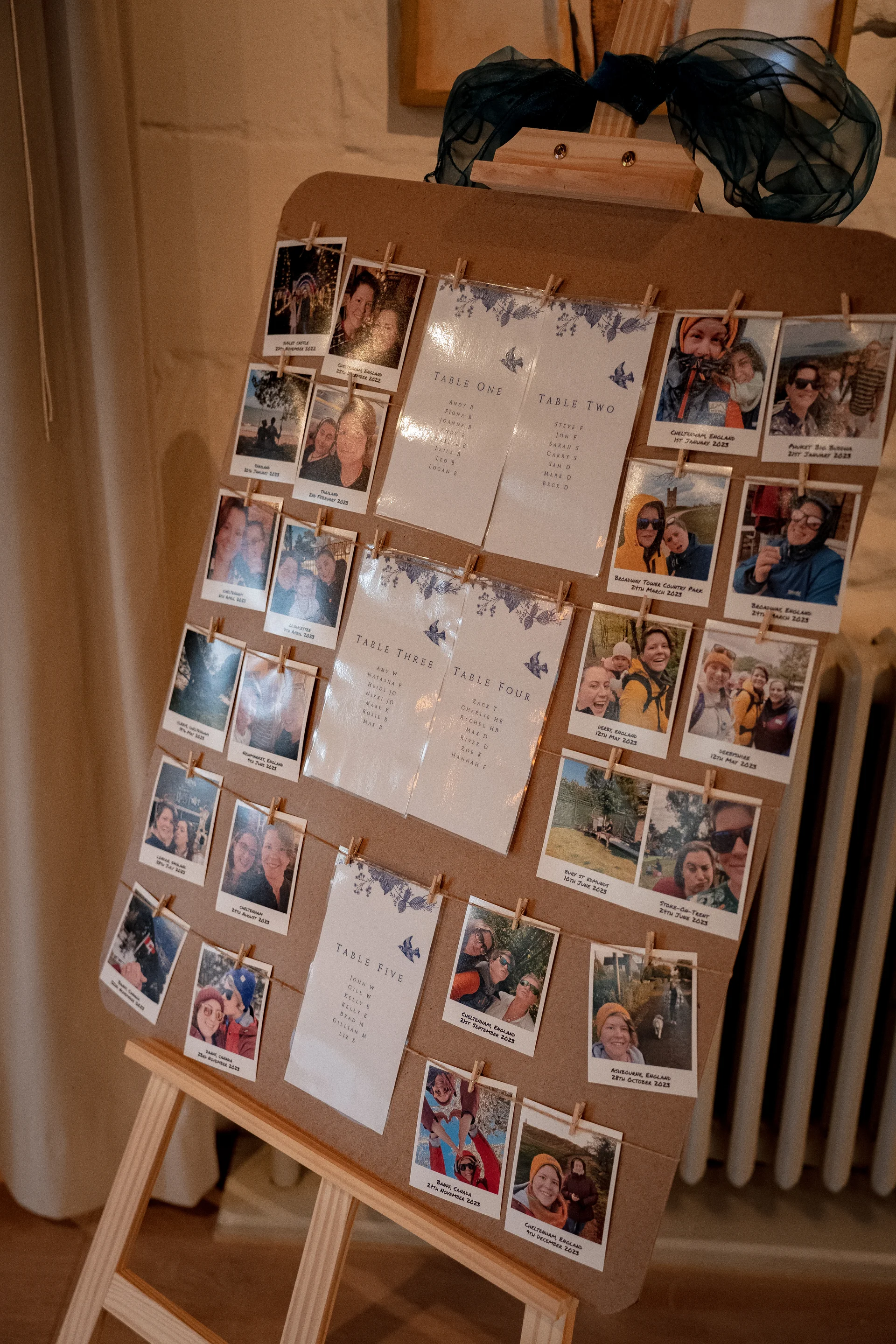 A rustic wooden easel holds a wedding seating plan board decorated with printed table lists and pinned polaroid photographs.
