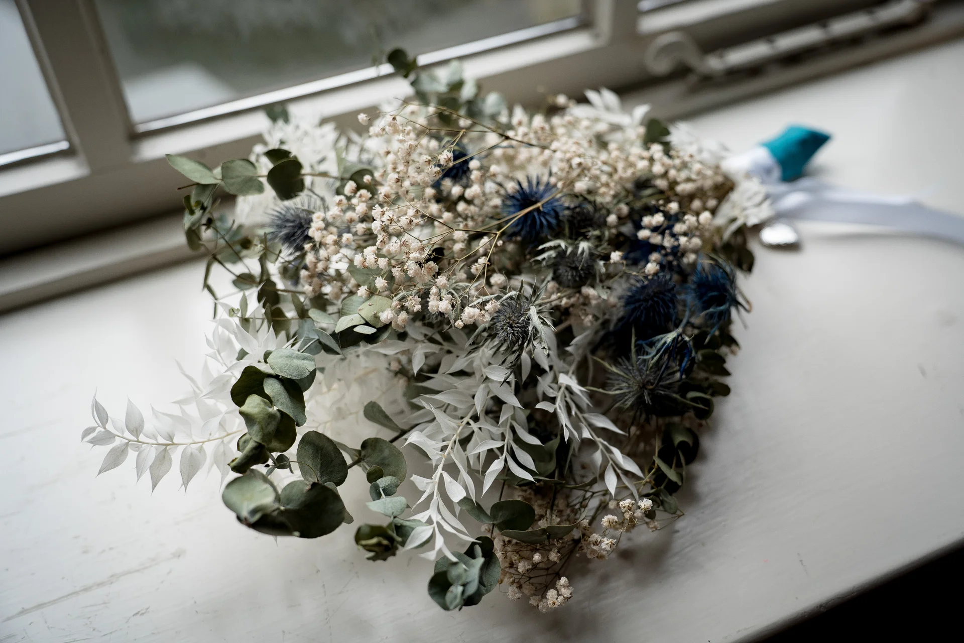 A detailed close-up of a dried bridal bouquet featuring thistles, eucalyptus, and baby's breath rests on a white windowsill bathed in natural window light.