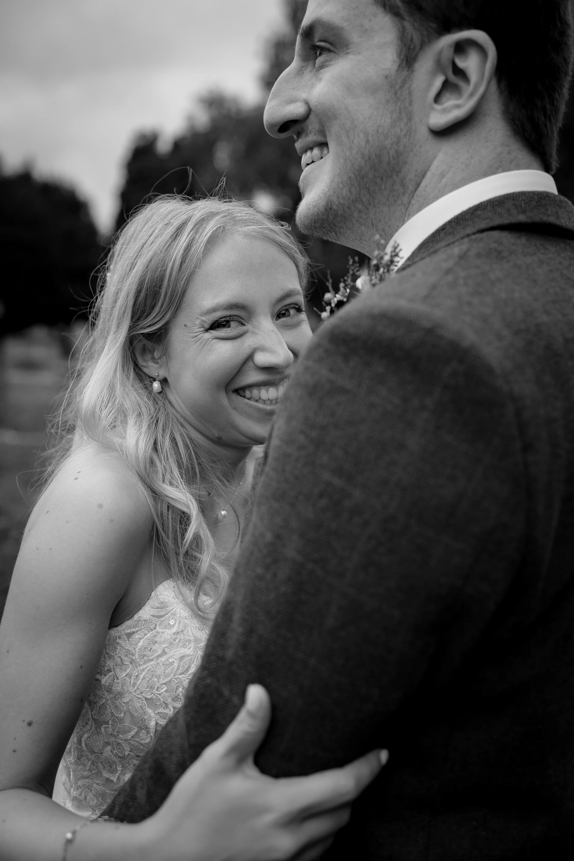 A smiling bride peeks over her groom's shoulder in an intimate, candid black and white portrait outdoors.