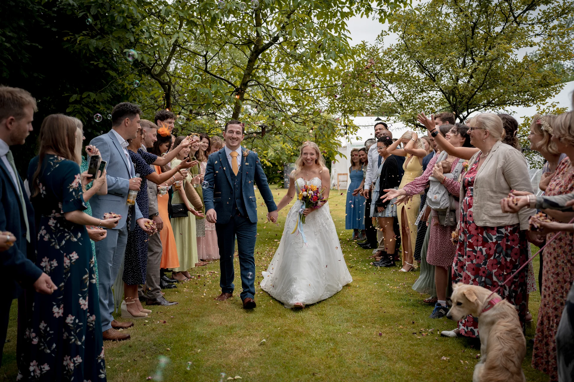 Newlyweds walk hand in hand down an outdoor aisle framed by cheering guests throwing confetti and blowing bubbles in a lush garden setting.