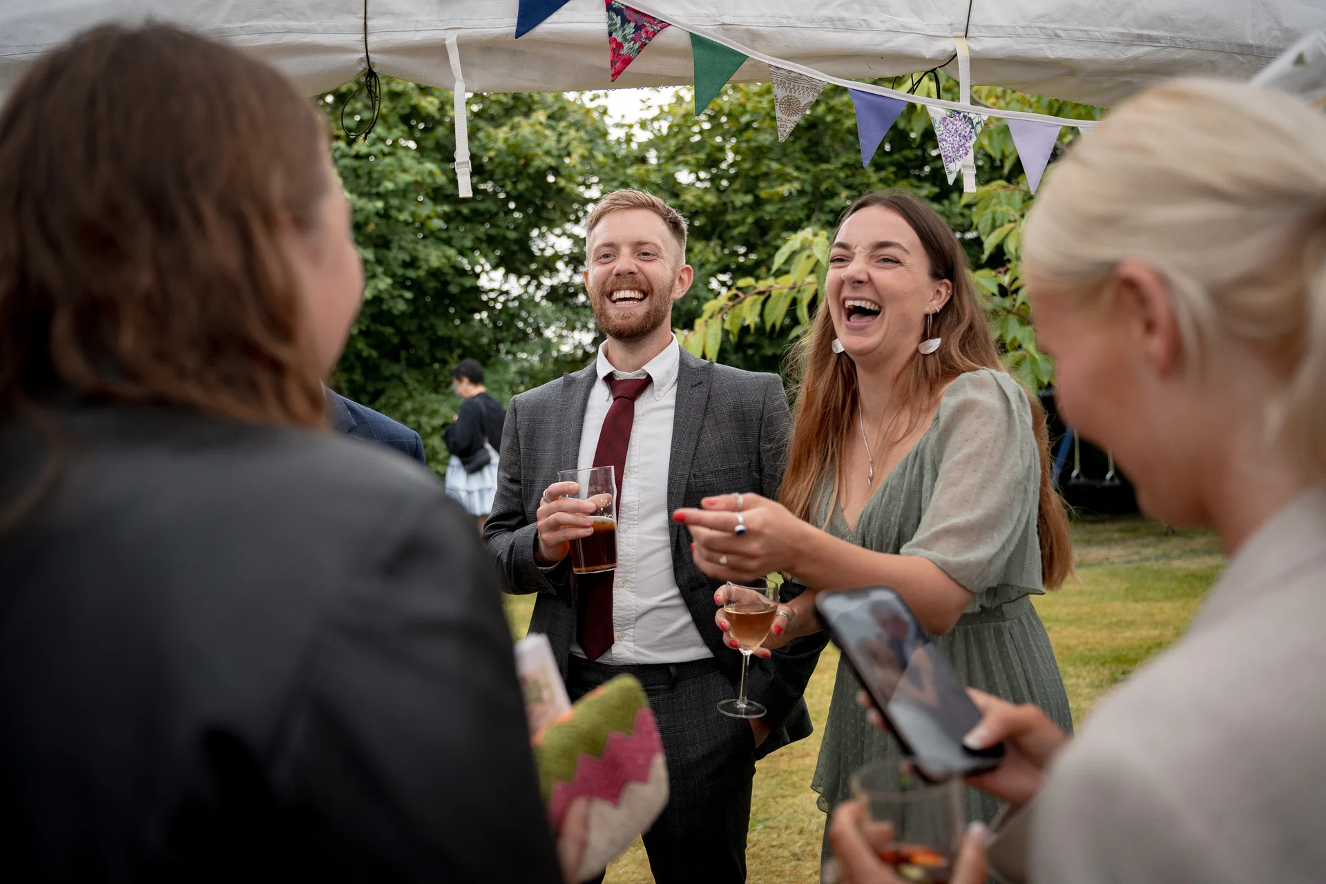 Wedding guests share a joyful, laughing moment together during a relaxed outdoor drinks reception under a marquee decorated with colourful bunting.